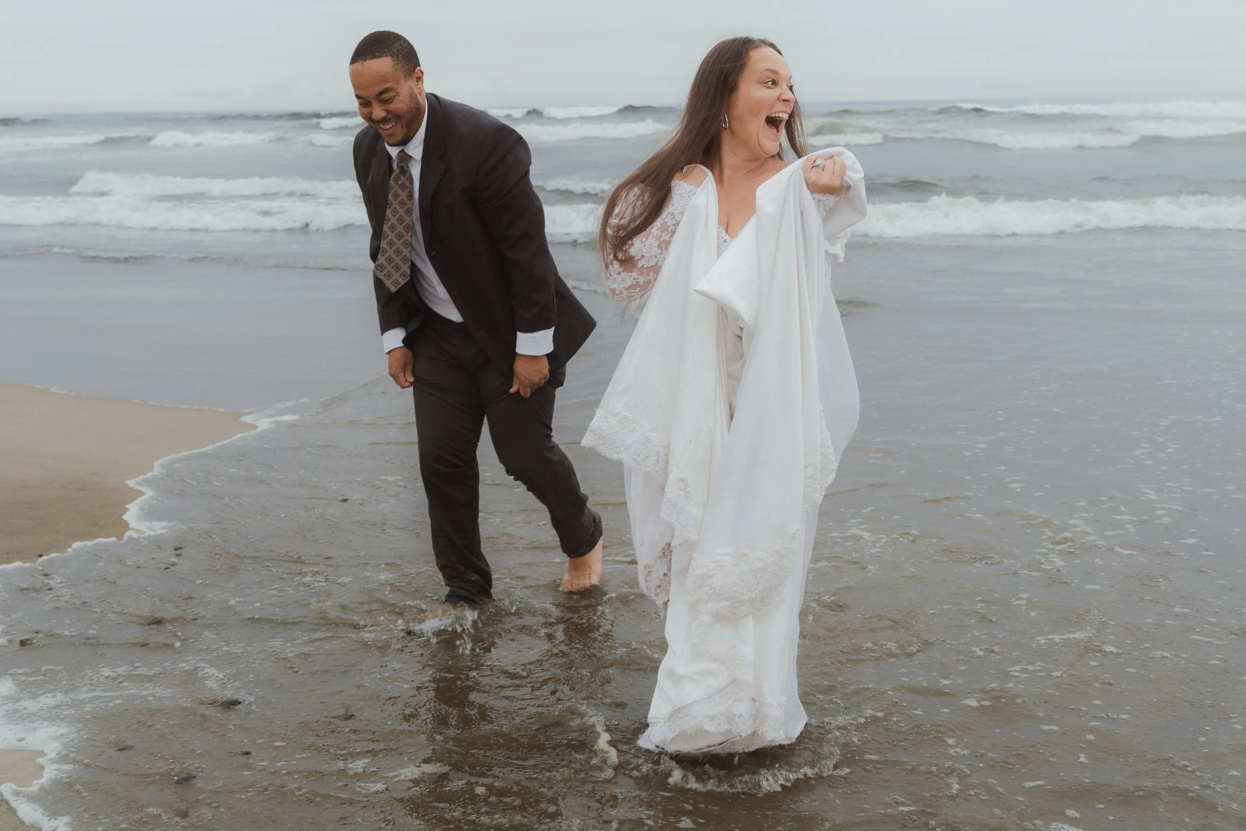 A happy couple dressed in wedding attire walking barefoot along the shoreline, with the man in a suit and the woman in a white wedding dress, both laughing as they enjoy the beach.
