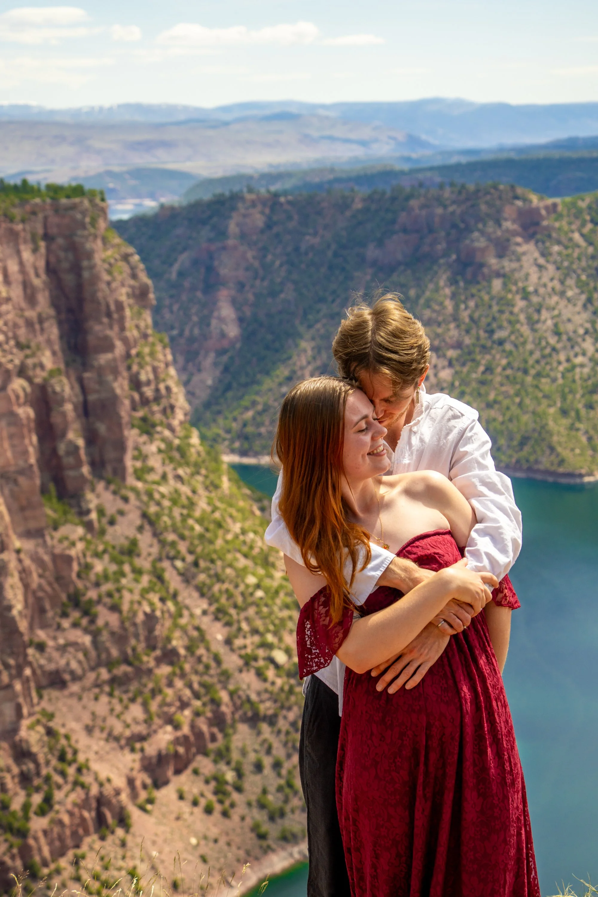 A couple hugging and kissing in front of a canyon and river landscape with mountains in the background.