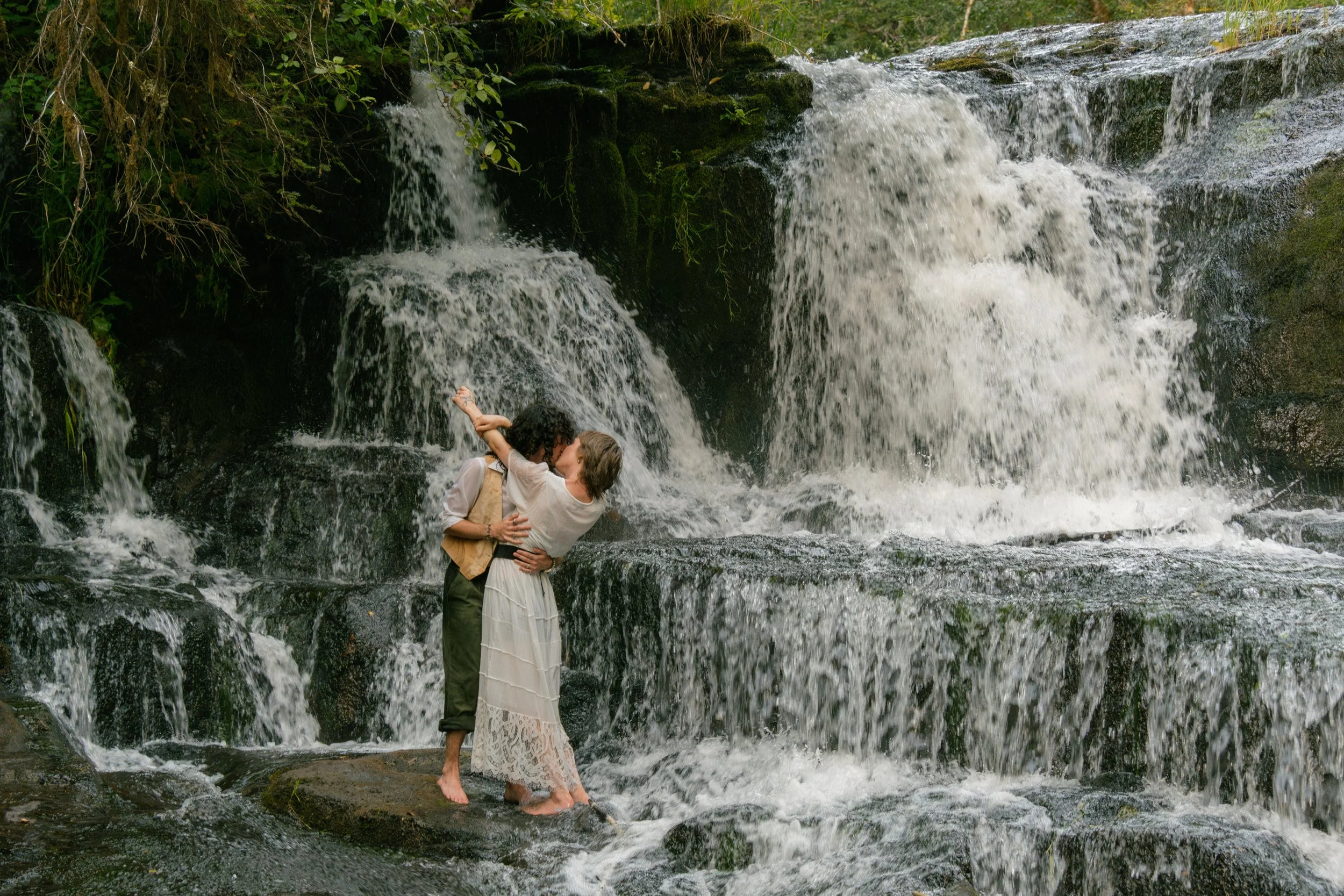 A couple standing in a rushing stream with a small waterfall behind them, embracing and about to kiss amid lush green foliage.