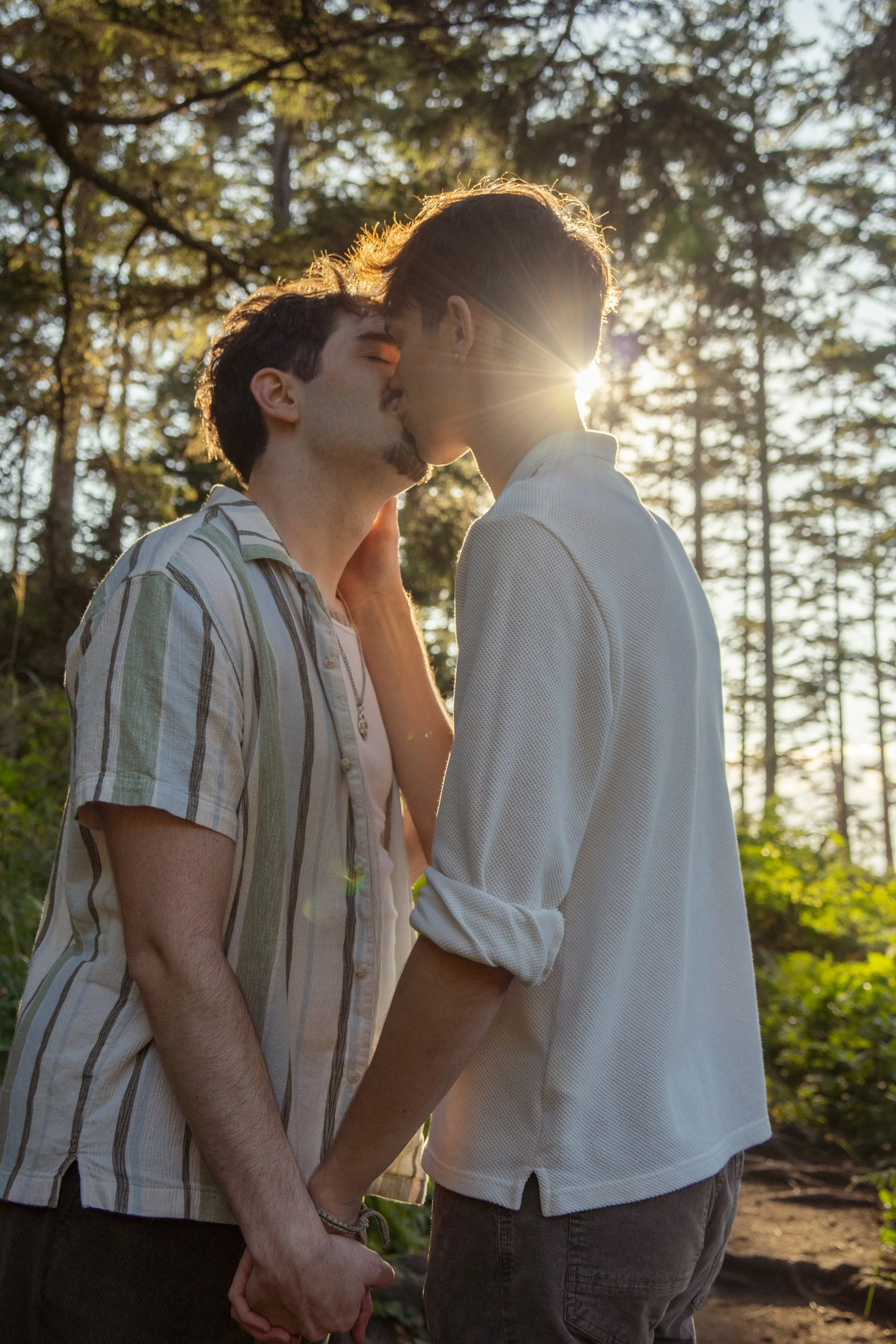 A couple kissing outdoors during sunset in a wooded area, holding hands with trees in the background.