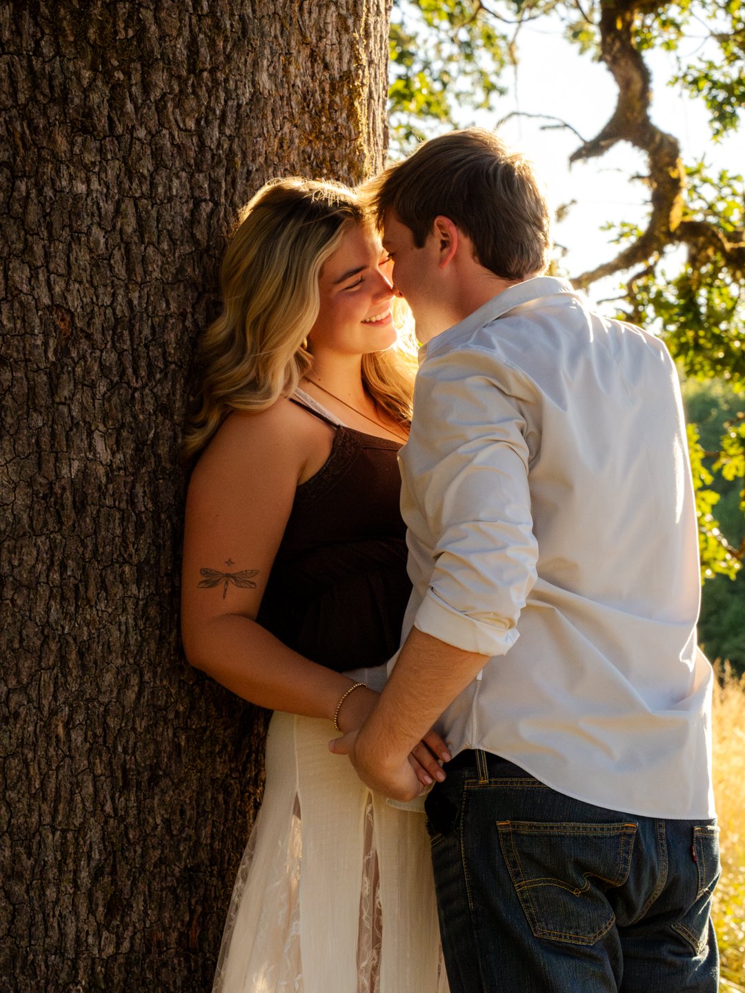 A young couple is leaning against a tree, smiling and touching foreheads, in a warm outdoor setting during sunset.