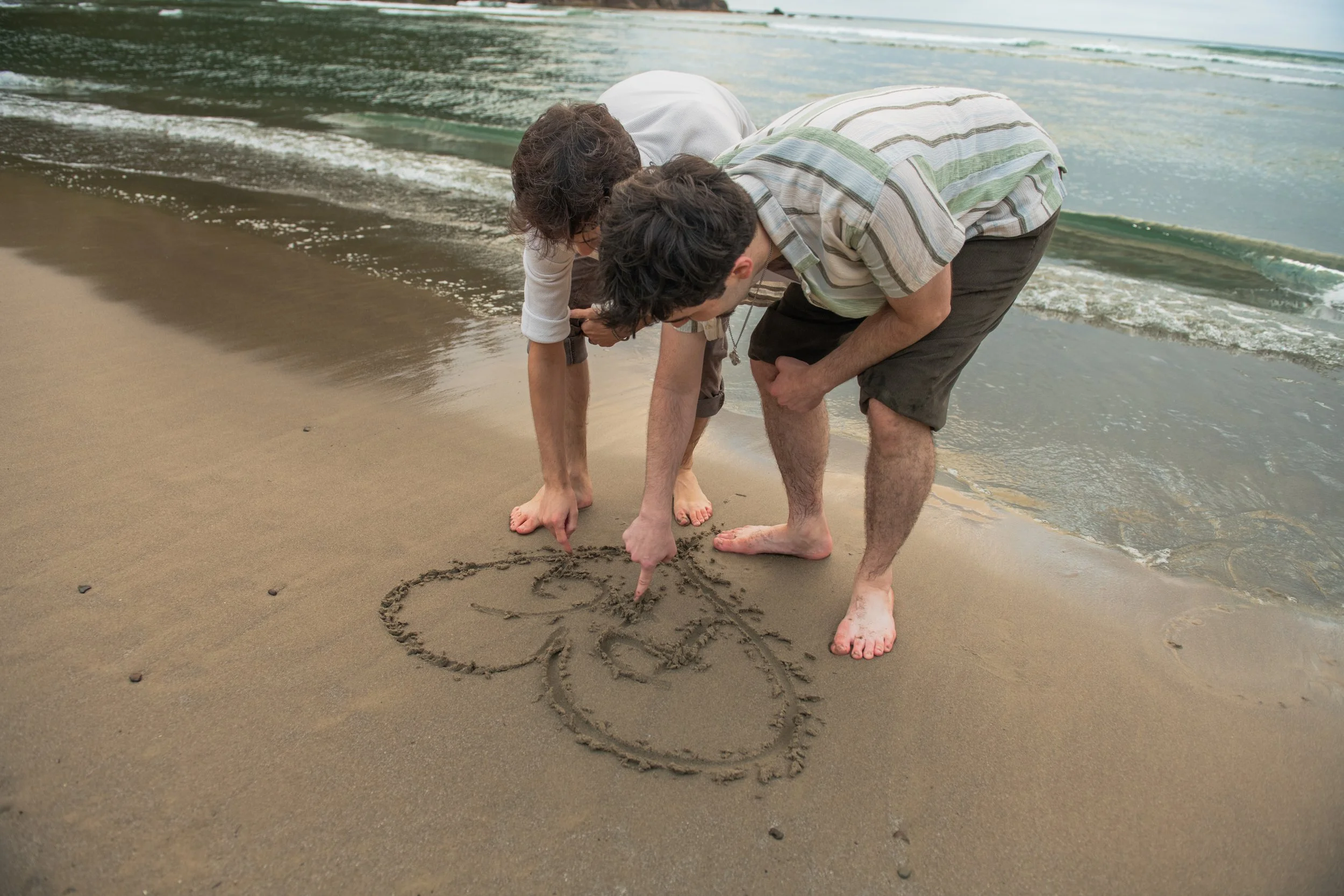 Two men drawing a heart in the sand at the beach on the Oregon Coast, one person is bending down and the other is pointing to the drawing.