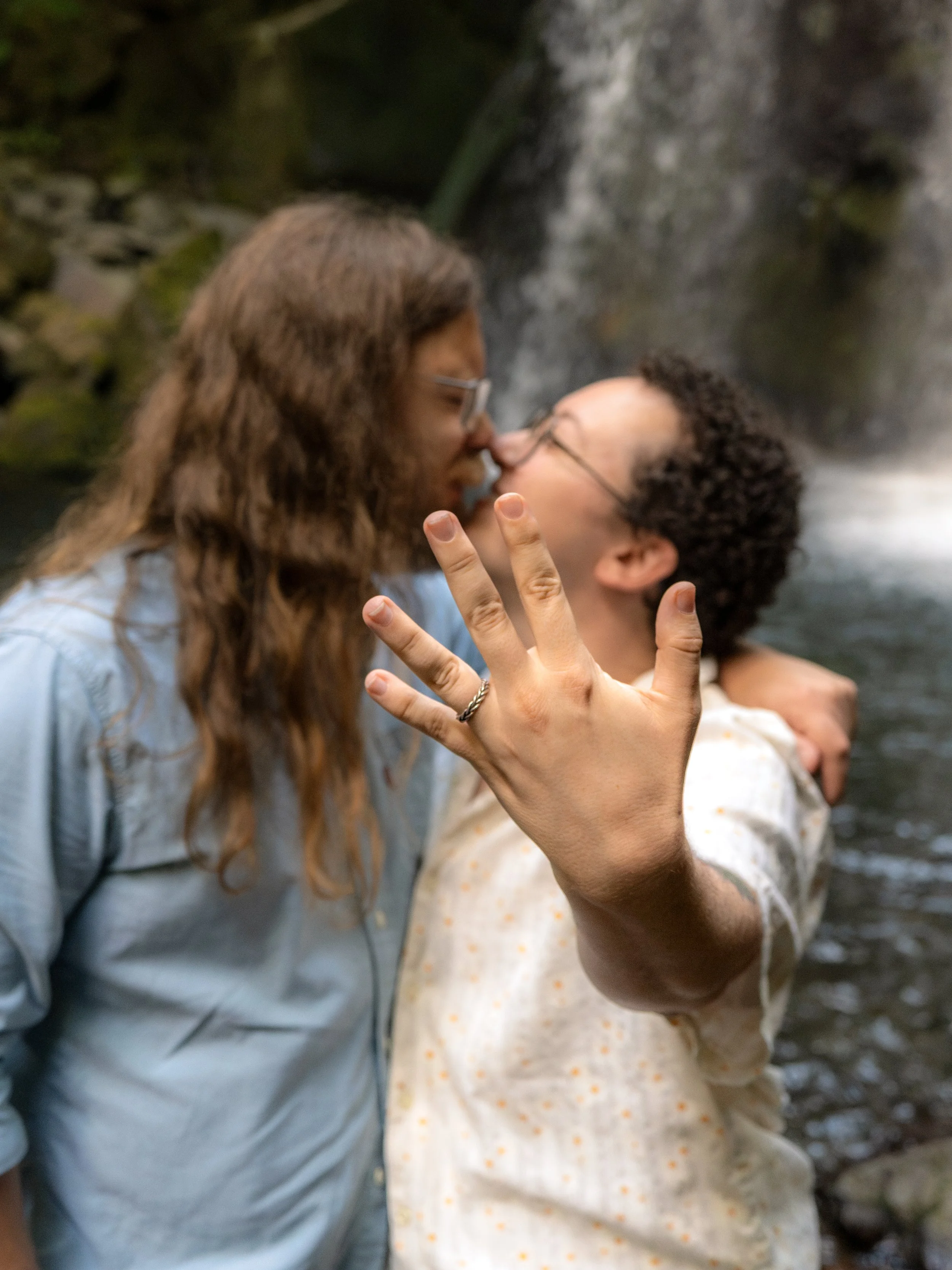a couple kissing showing an engagement ring in front of a waterfall
