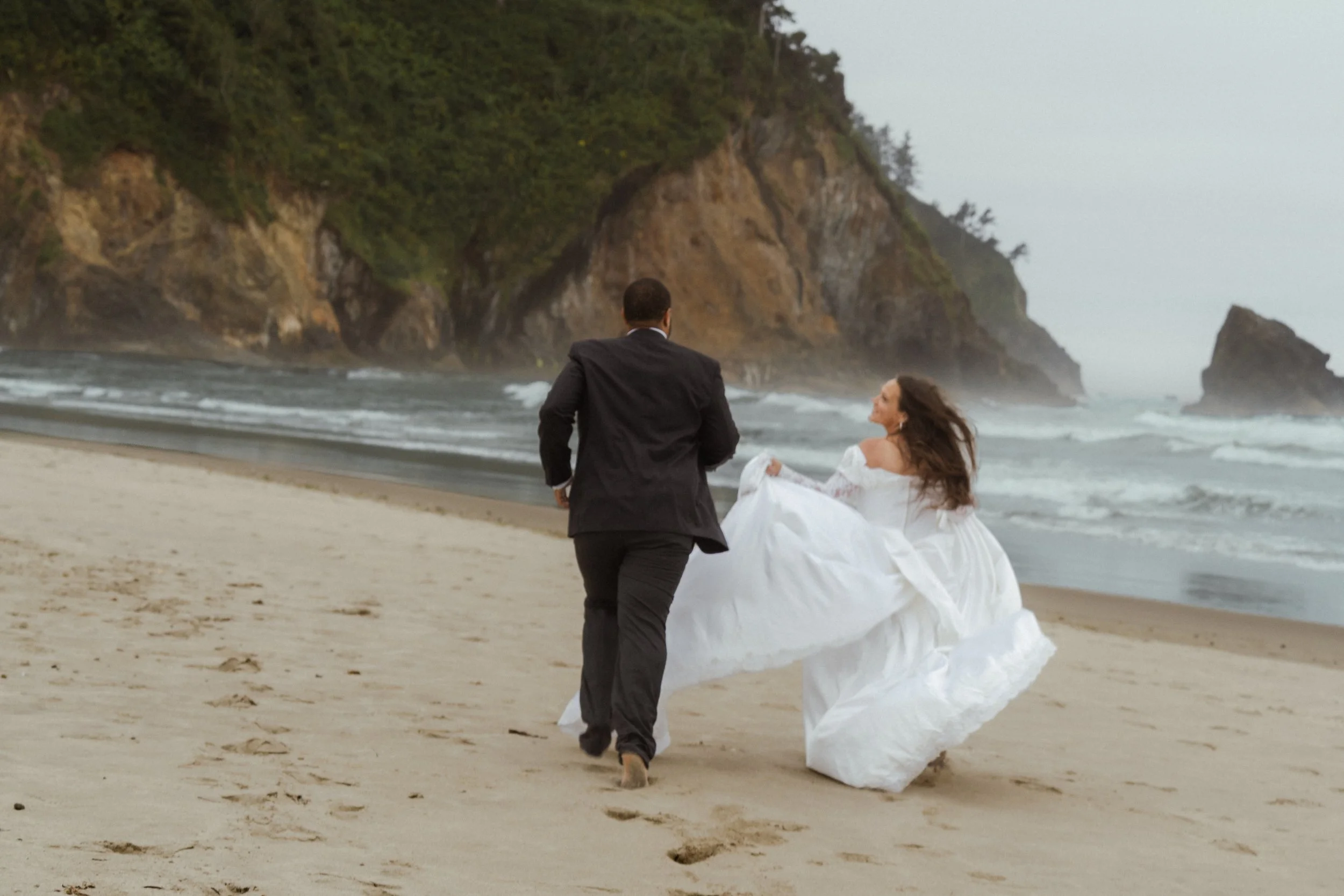 A man in a dark suit walking barefoot on the beach with a woman in a white dress, the woman is sitting or lying down close to the shoreline, with rocky cliffs and trees in the background.