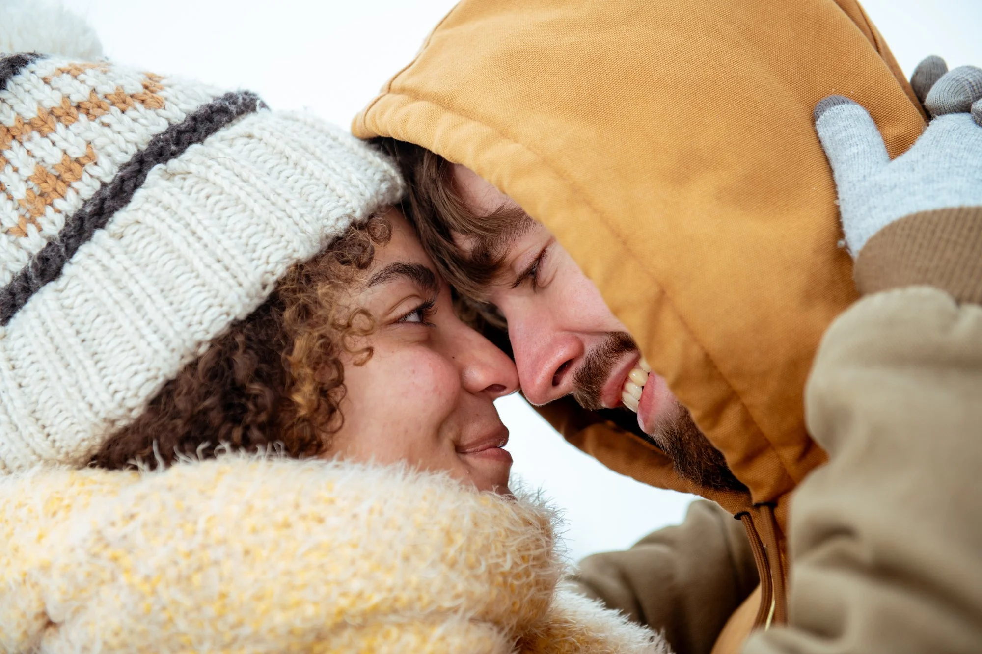 Close-up of a young woman with curly hair and a young man with brown hair and a beard, both smiling and touching foreheads, in warm winter clothing including a knitted hat, gloves, and jackets.