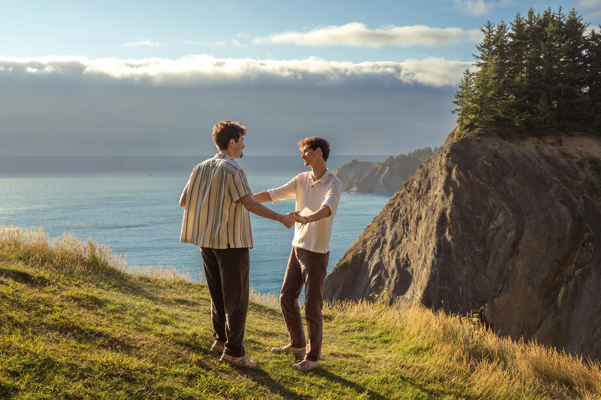 Two people holding hands and smiling at each other on a grassy cliffside with the ocean in the background, under a partly cloudy sky.