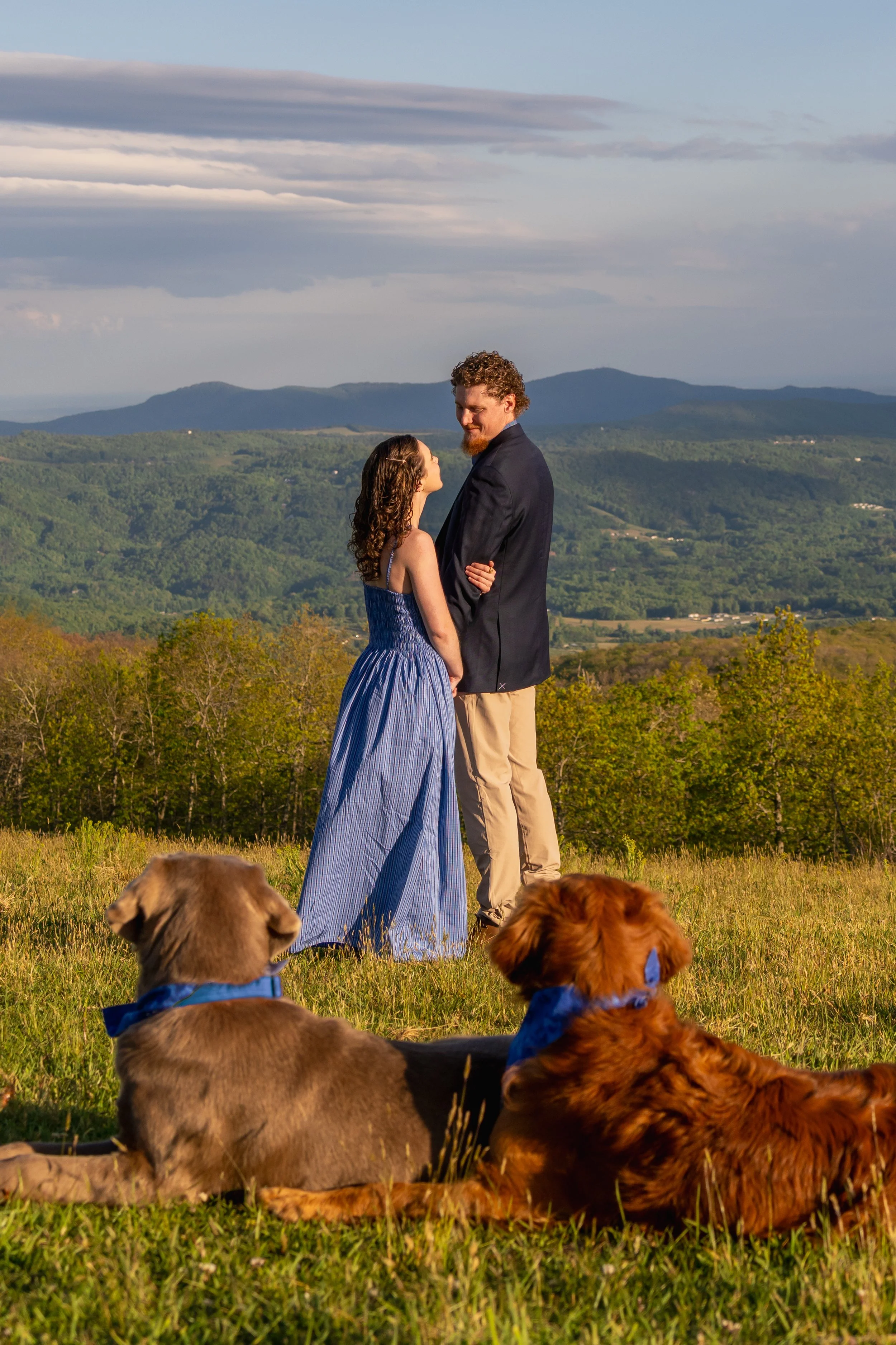 A couple standing on a grassy hilltop with scenic mountains in the background, surrounded by three dogs, during daylight.