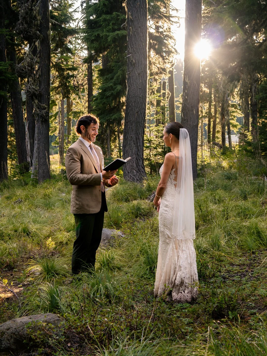 A wedding ceremony taking place in a forest with a groom reading vows and a bride listening, both dressed in formal attire with the bride in a white lace gown and veil, sunlight shining through tall trees.