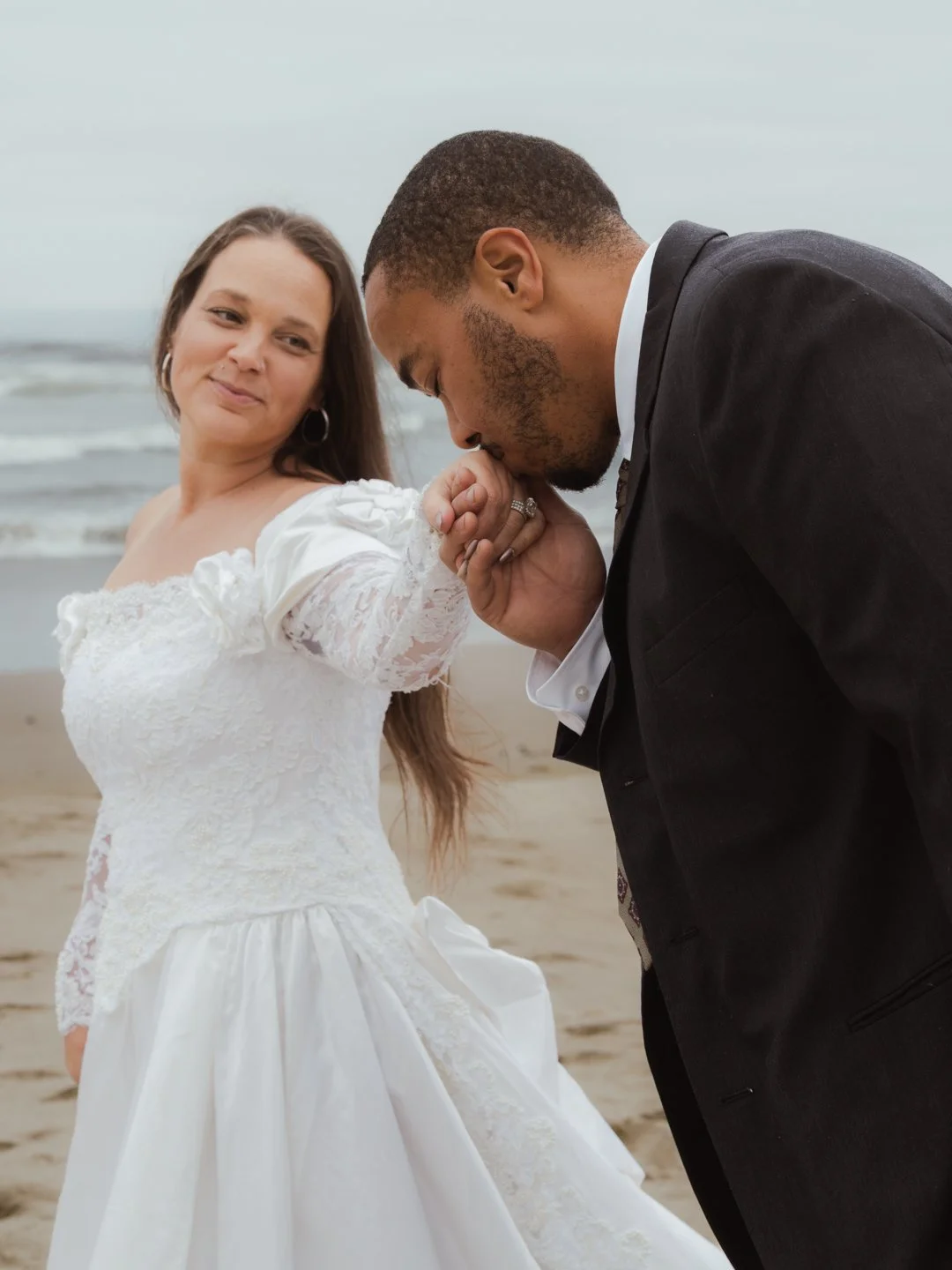 A bride in a white lace wedding dress holds hands with a groom in a black suit who is kissing her hand on a beach.