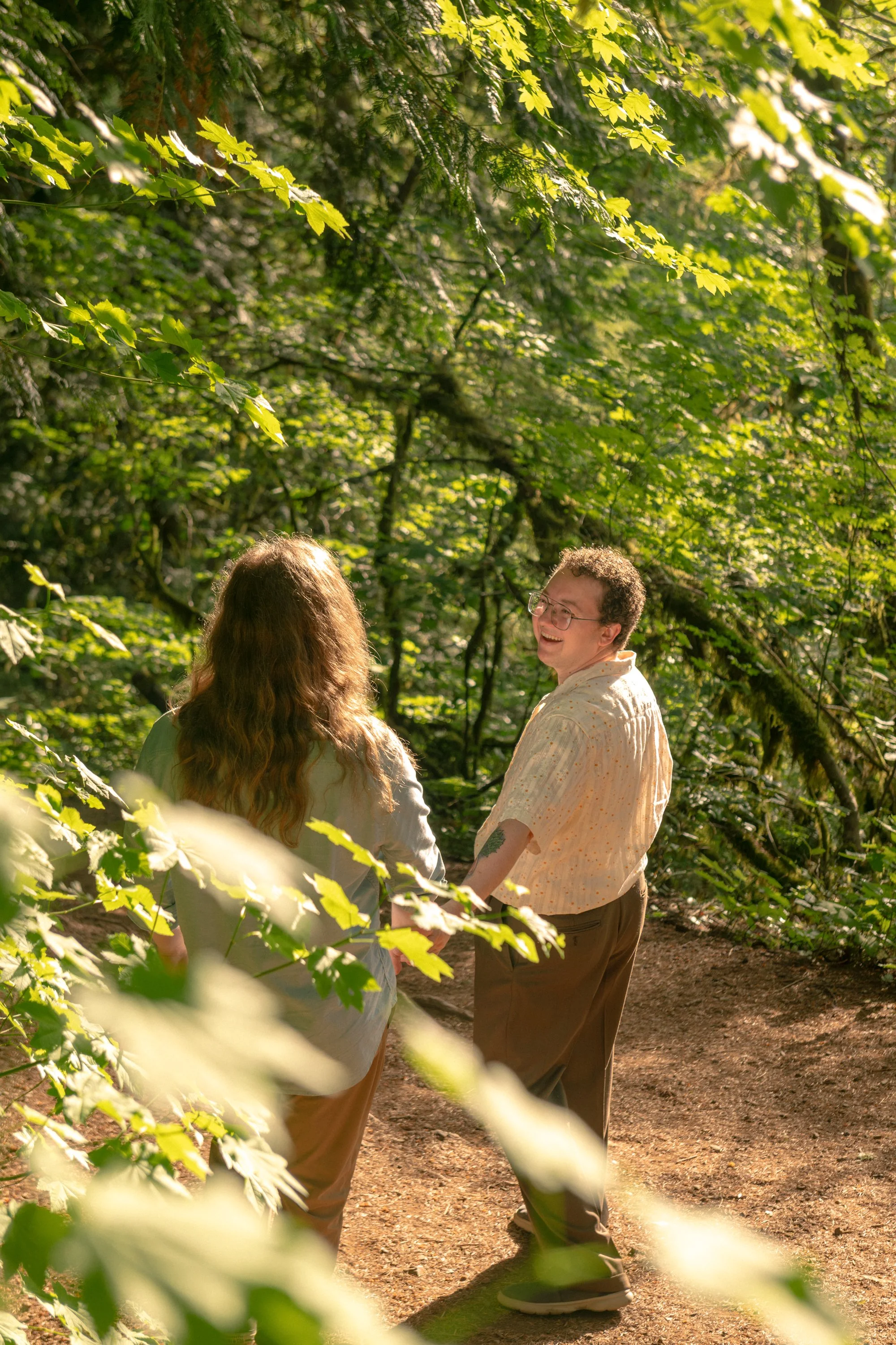 Two people walking along a wooded forest trail, smiling and talking to each other, surrounded by green foliage and sunlight filtering through the trees.