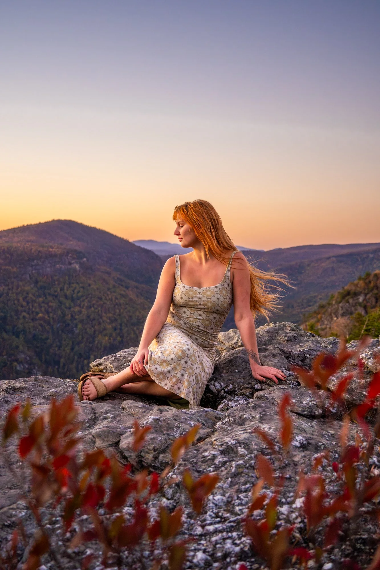 A woman with red hair sitting on rocks outdoors during sunset in a mountainous area, wearing a patterned dress and sandals, with red foliage in the foreground.