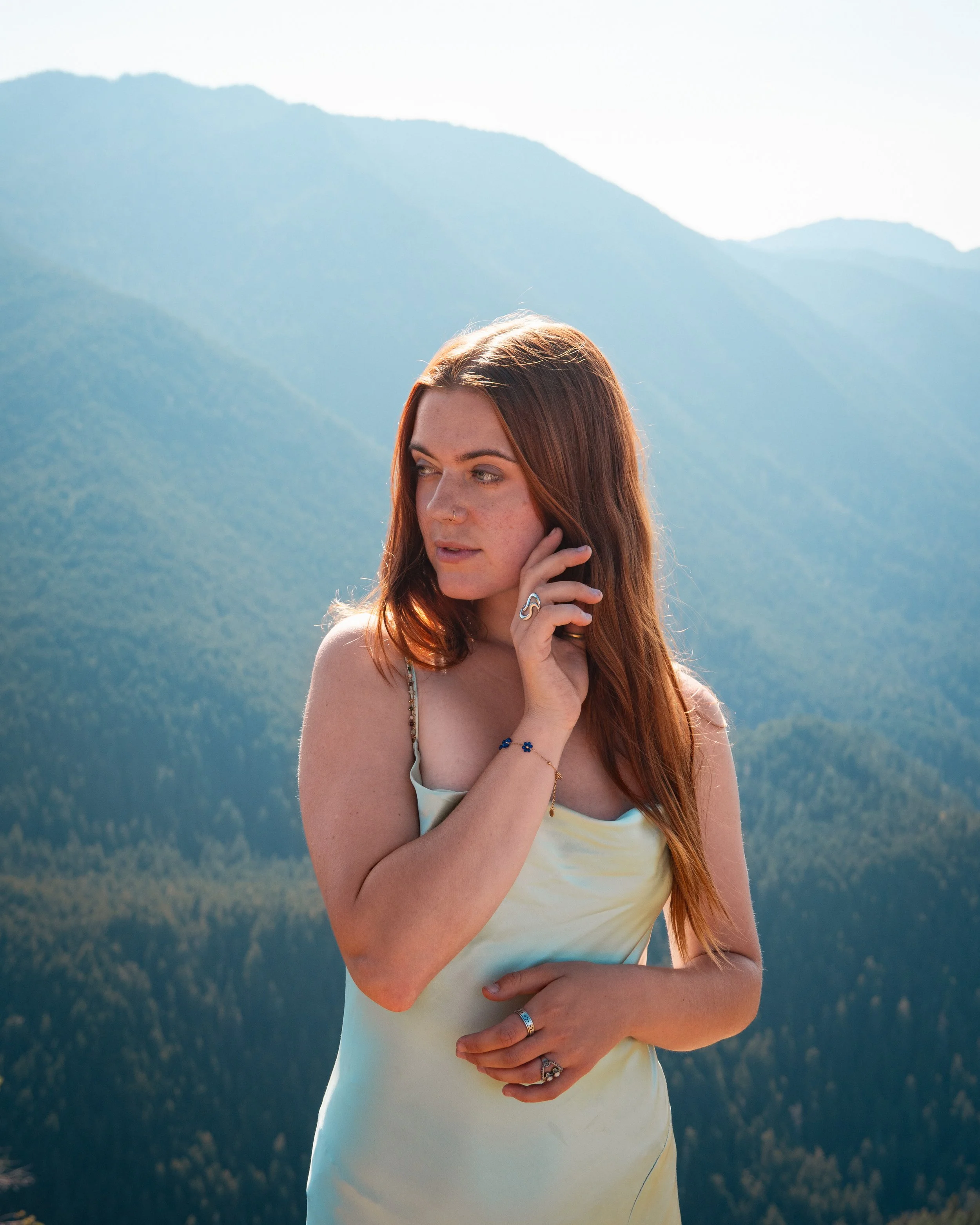 A woman with red hair and fair skin standing outdoors against a backdrop of the Olympic Mountains. She is wearing a blue dress and jewelery. Her right hand touches her face, and her left hand is at her waist.