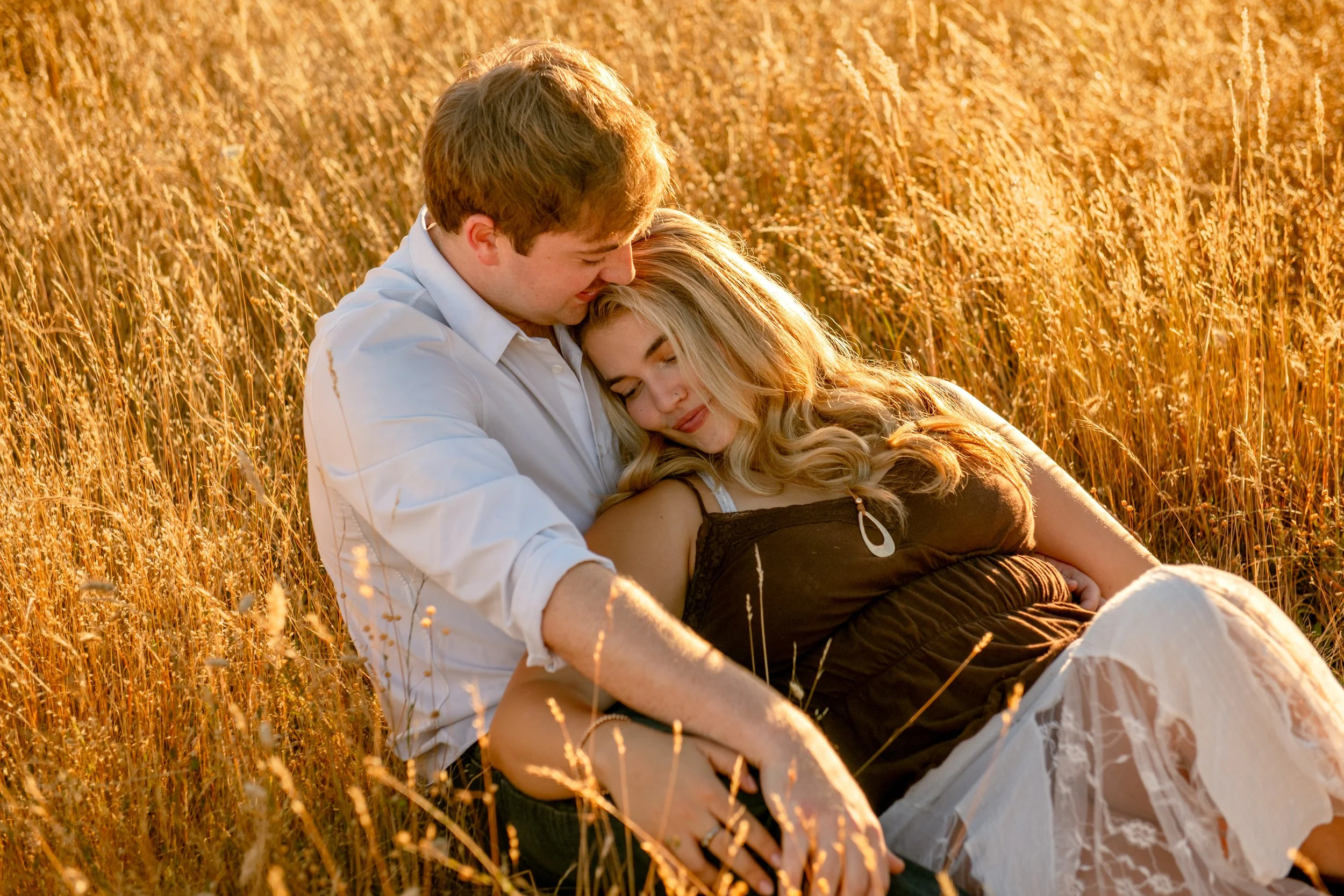 A young couple sitting in a wheat field at sunset, cuddling and smiling peacefully.