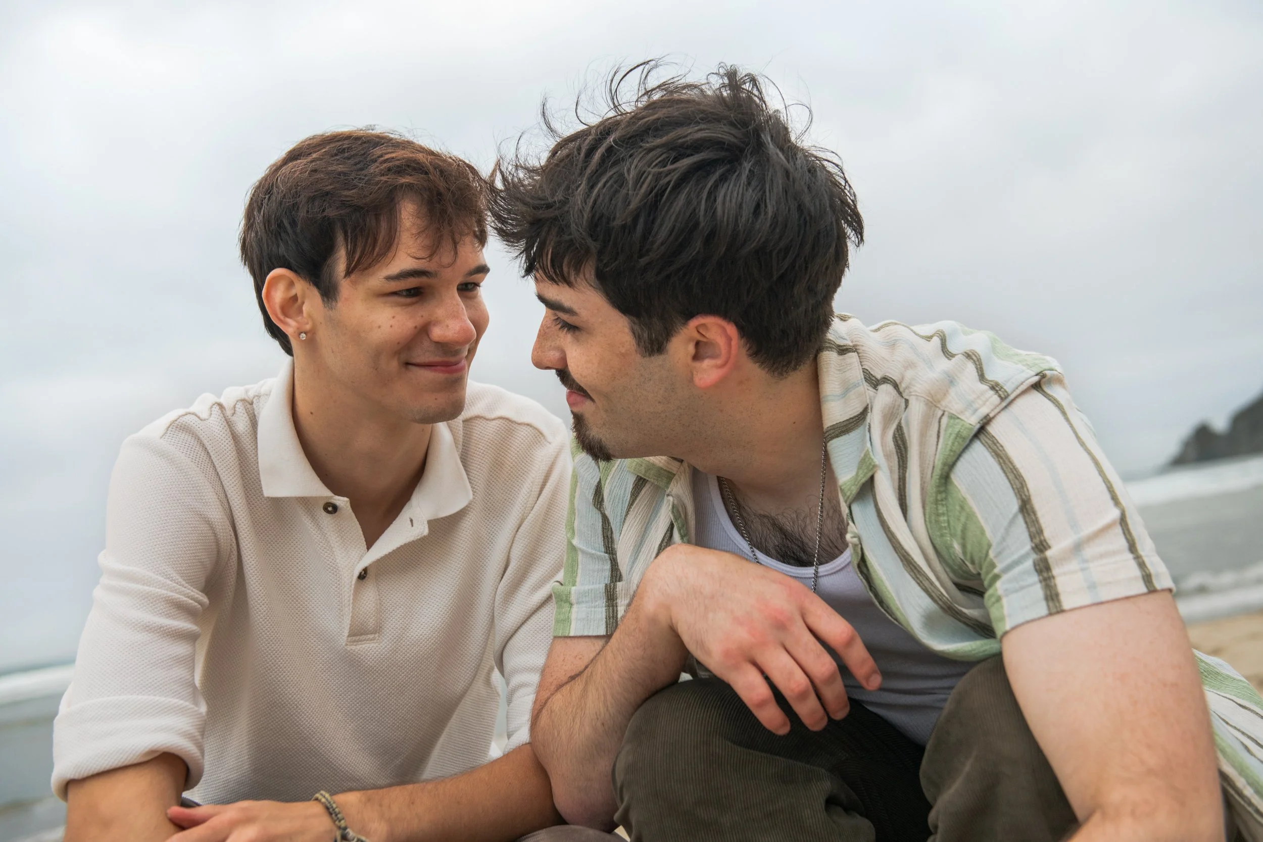 Two men sit close together outdoors, facing each other, with gentle, smiling expressions, against a cloudy sky background.