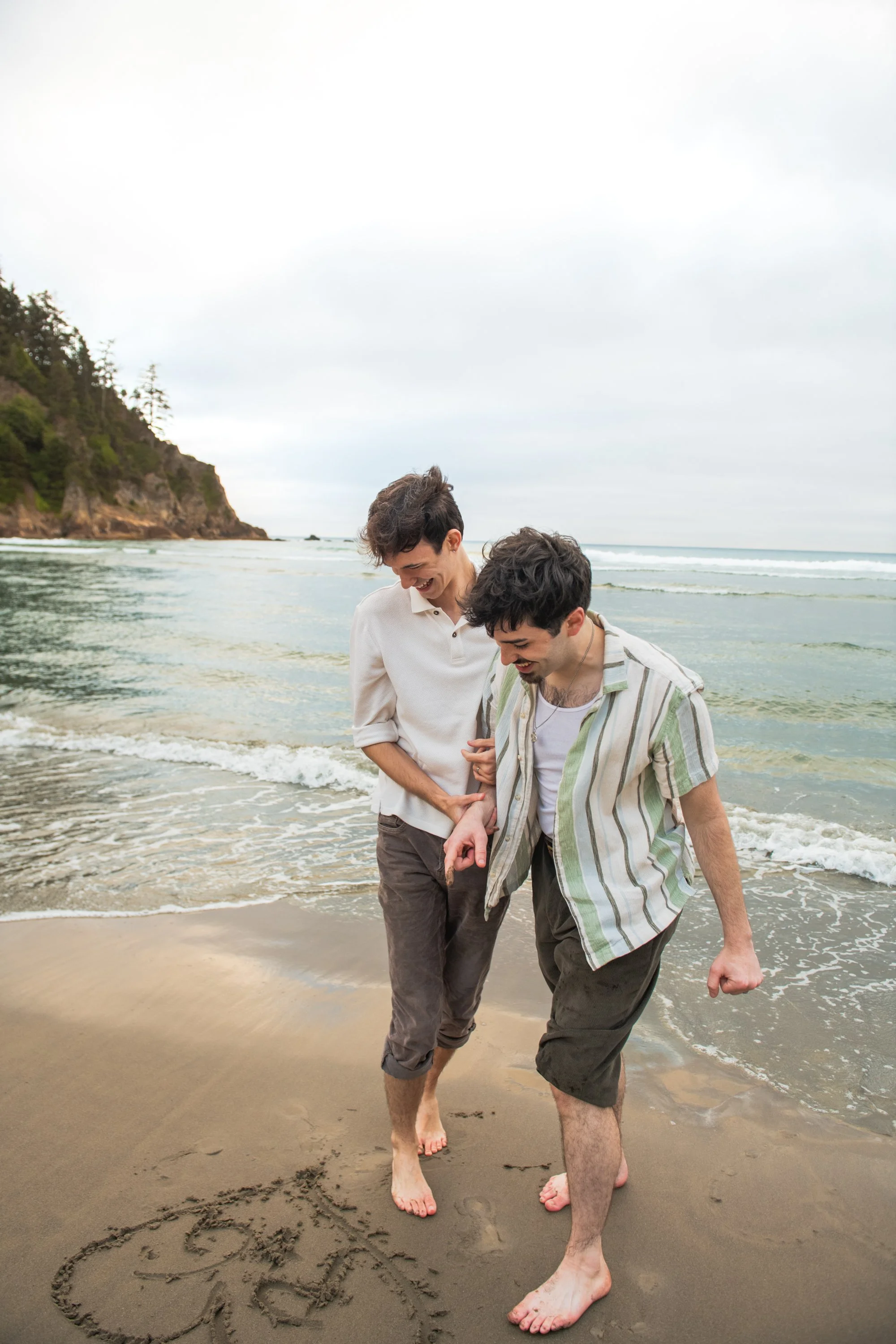 Two young men looking at the heart they drew in the sand on the Oregon coast, smiling and holding hands.