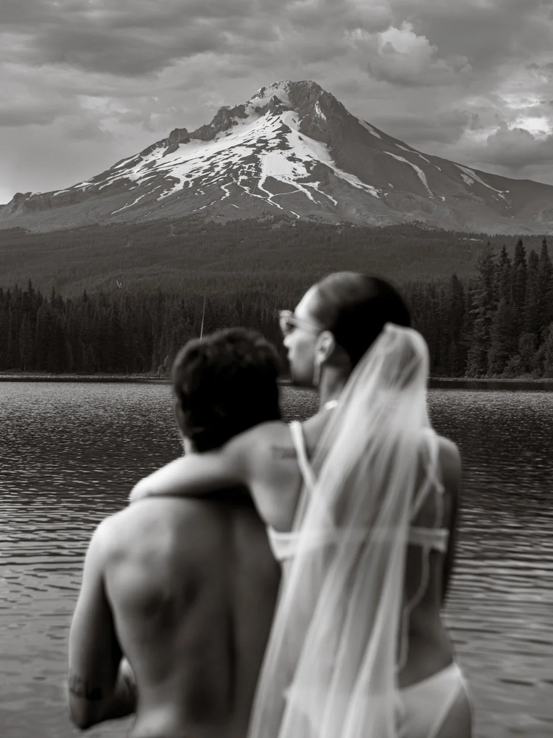 A black and white photo of a couple in wedding attire embracing near a lake, with a mountain and forest in the background.
