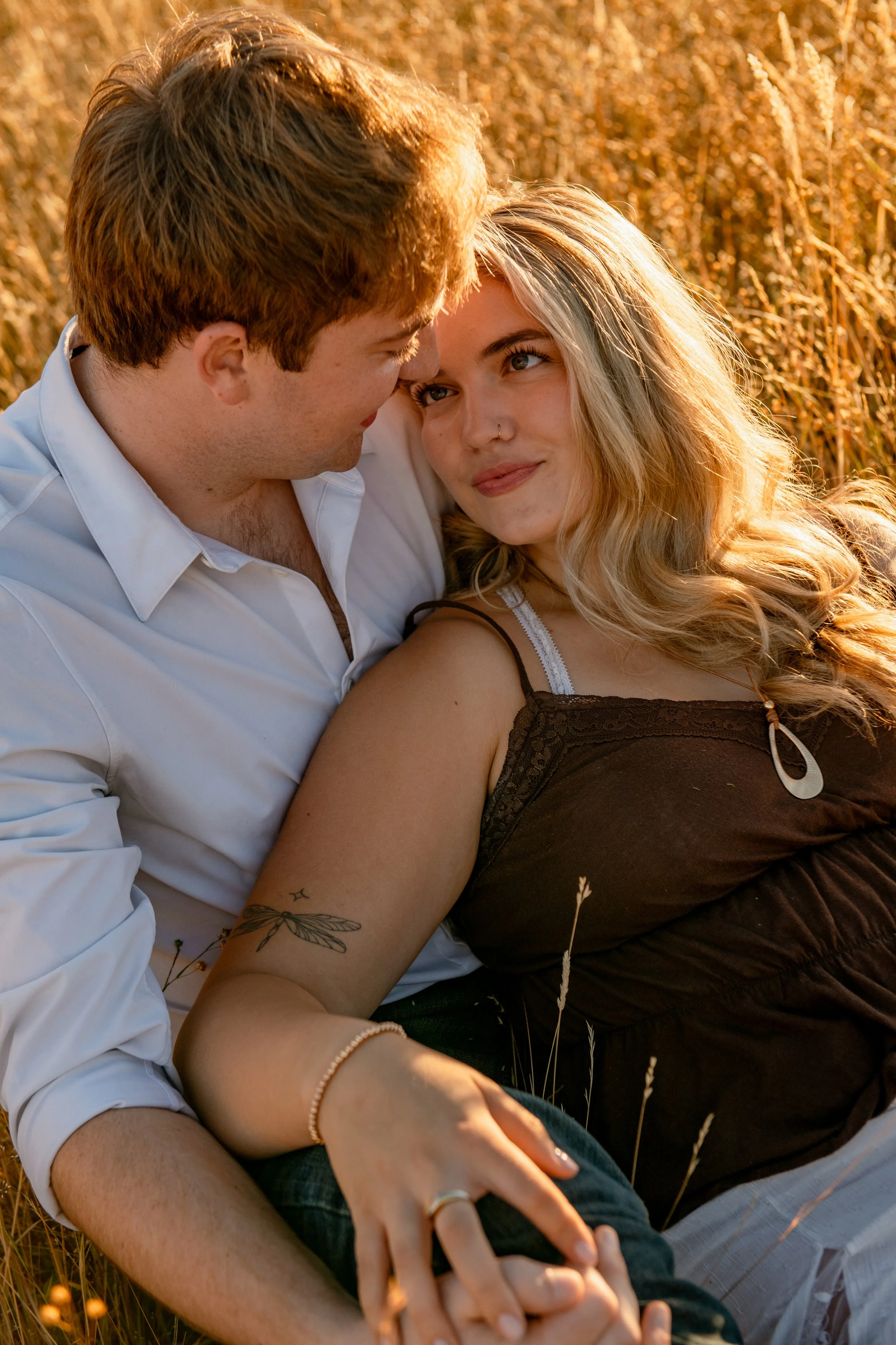 A couple lying in a field of tall, golden grass during sunset, gazing lovingly at each other.