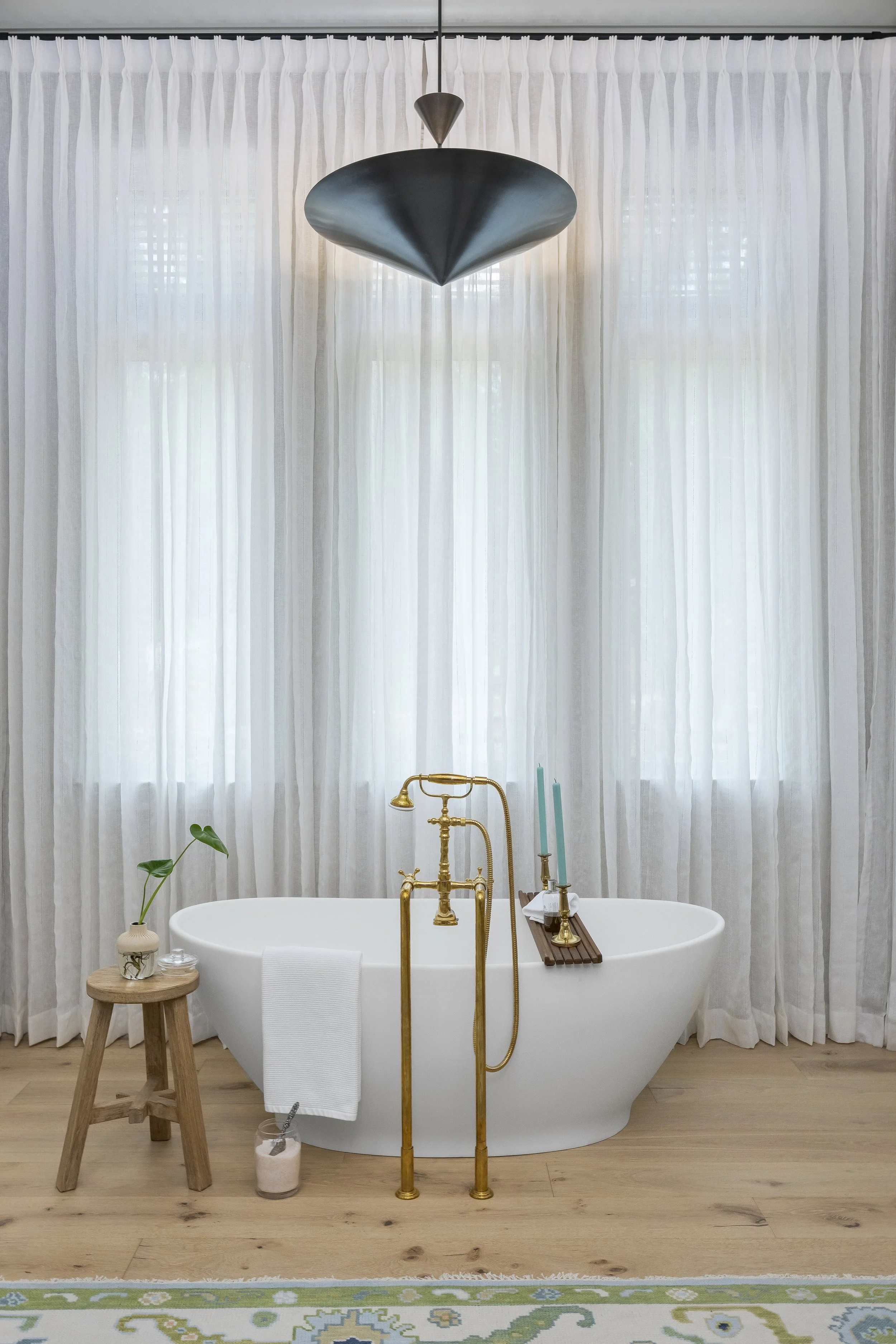 A modern bathroom with a white freestanding bathtub, gold fixtures, and sheer white curtains. A small wooden stool holds a potted plant, and candles are on the bathtub.