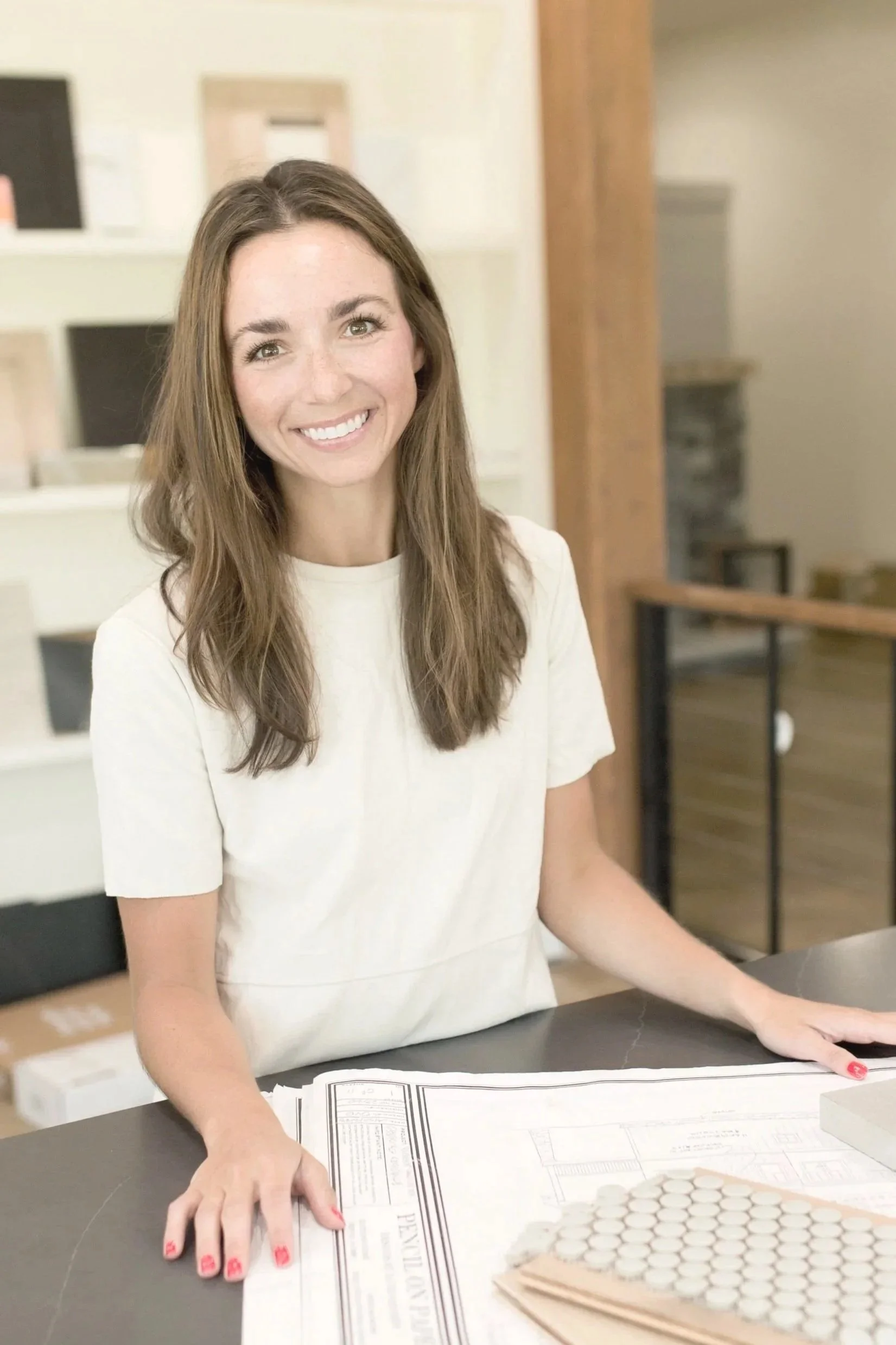 A woman with long brown hair and a white shirt smiling at a desk with architectural plans and color samples.