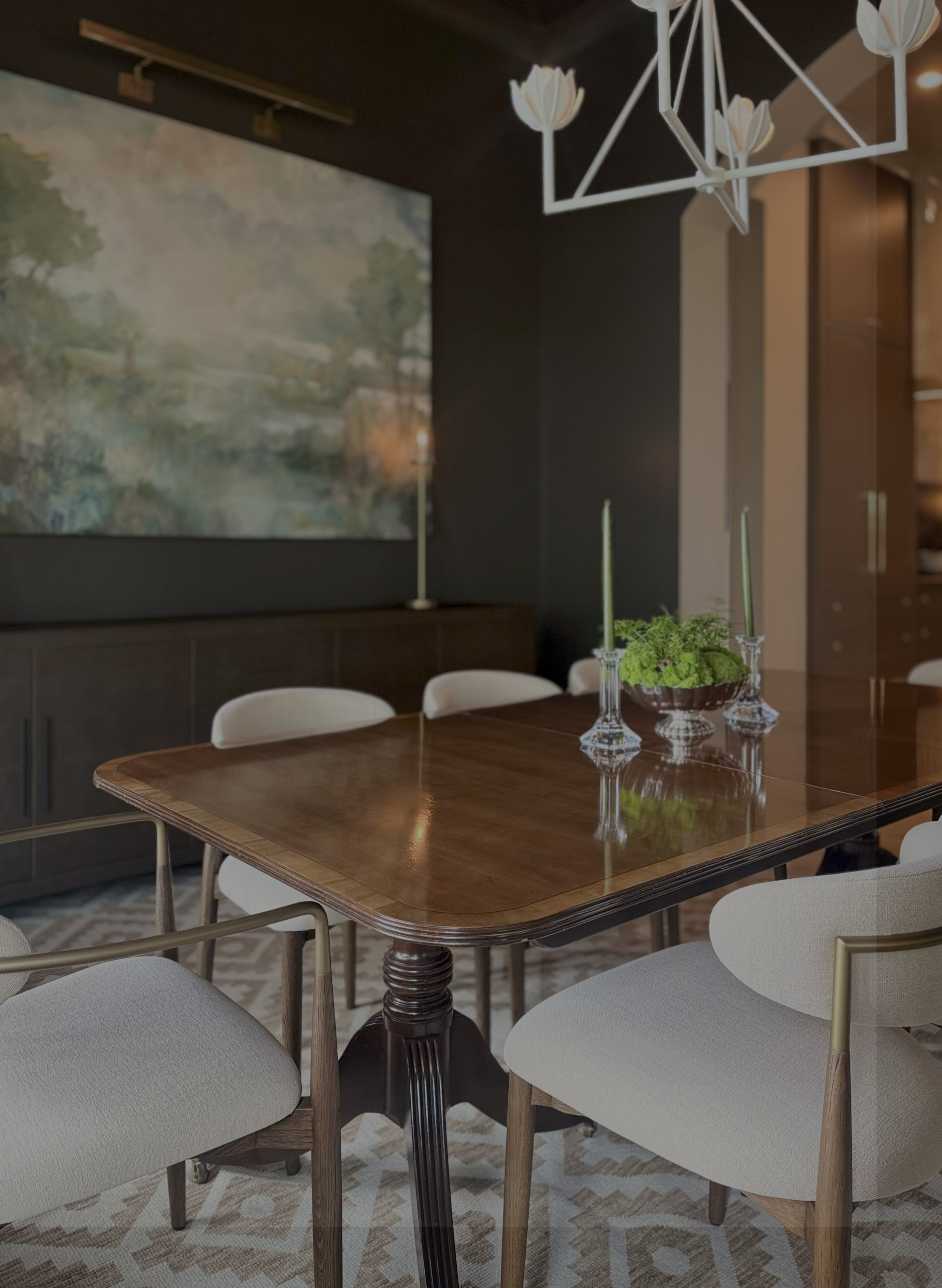 A dining room with a wooden table, white cushioned chairs, a modern geometric chandelier, and a large abstract landscape painting on a dark wall.