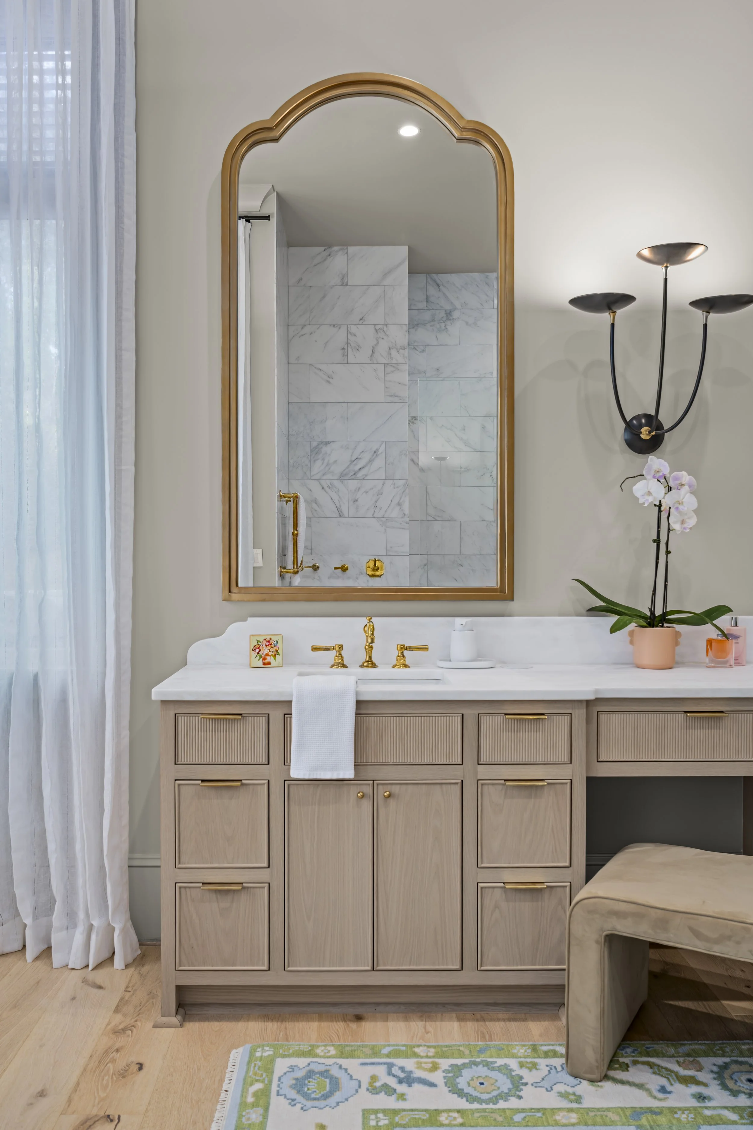 Bathroom vanity with a large mirror, beige cabinets, gold fixtures, orchid plant, and a painted stool. The mirror reflects a marble shower with gold fixtures, and white sheer curtains are seen on the side.