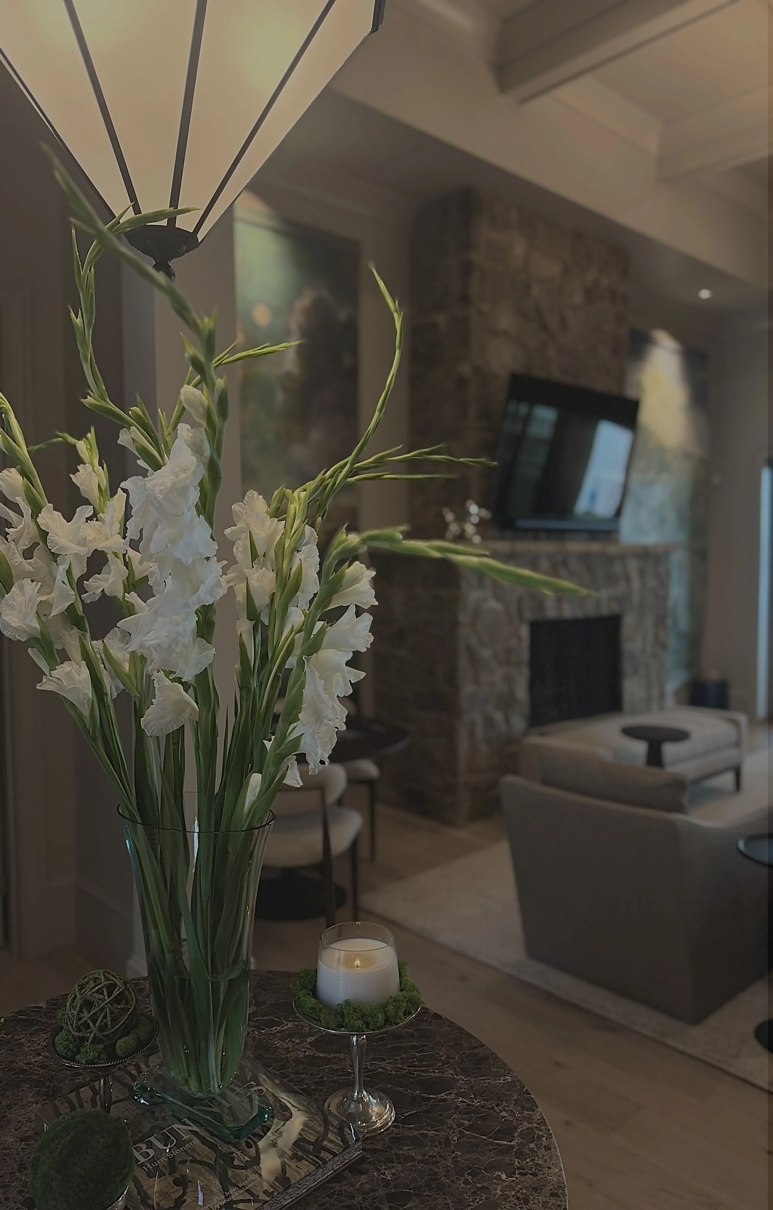A close-up view of a glass vase with white flowers on a marble table in a living room, with a stone fireplace and a mounted TV in the background.