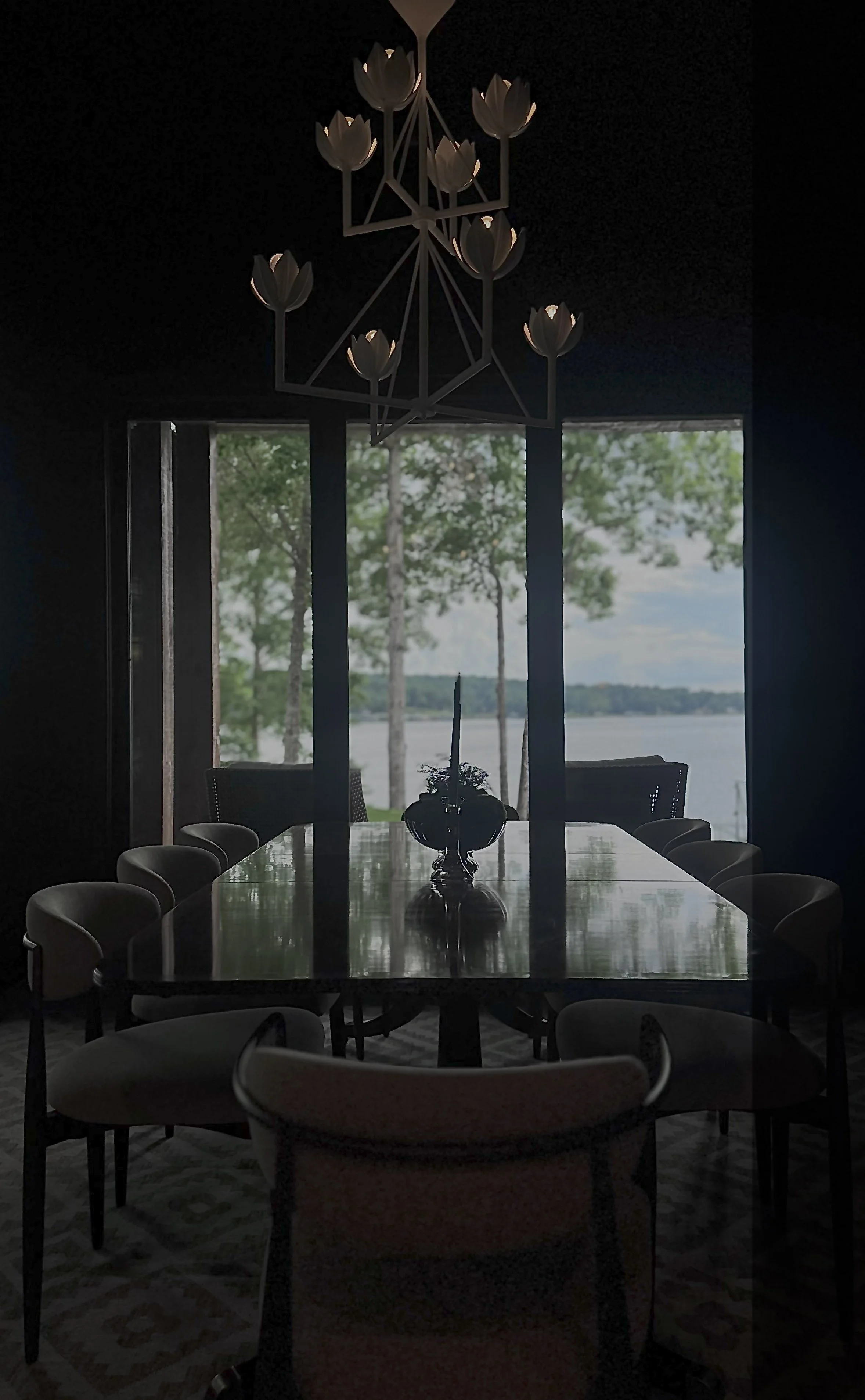 A dimly lit dining room with a large table, surrounded by chairs, and a dark chandelier with tulip-shaped lights hanging above. Large windows in the background reveal a lakeside view with trees and water.