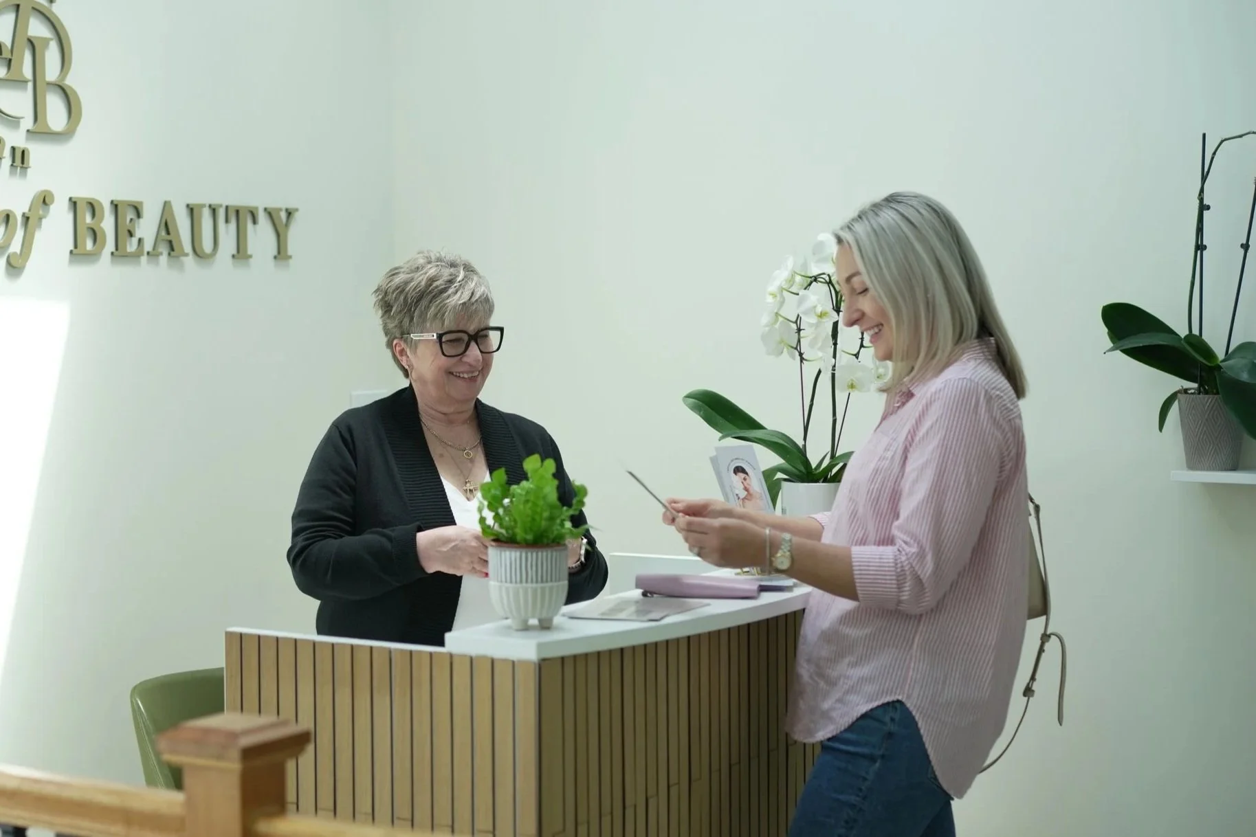 Women at a beauty clinic counter, one woman smiling at the receptionist, the other looking at her phone, with plants and decor around.