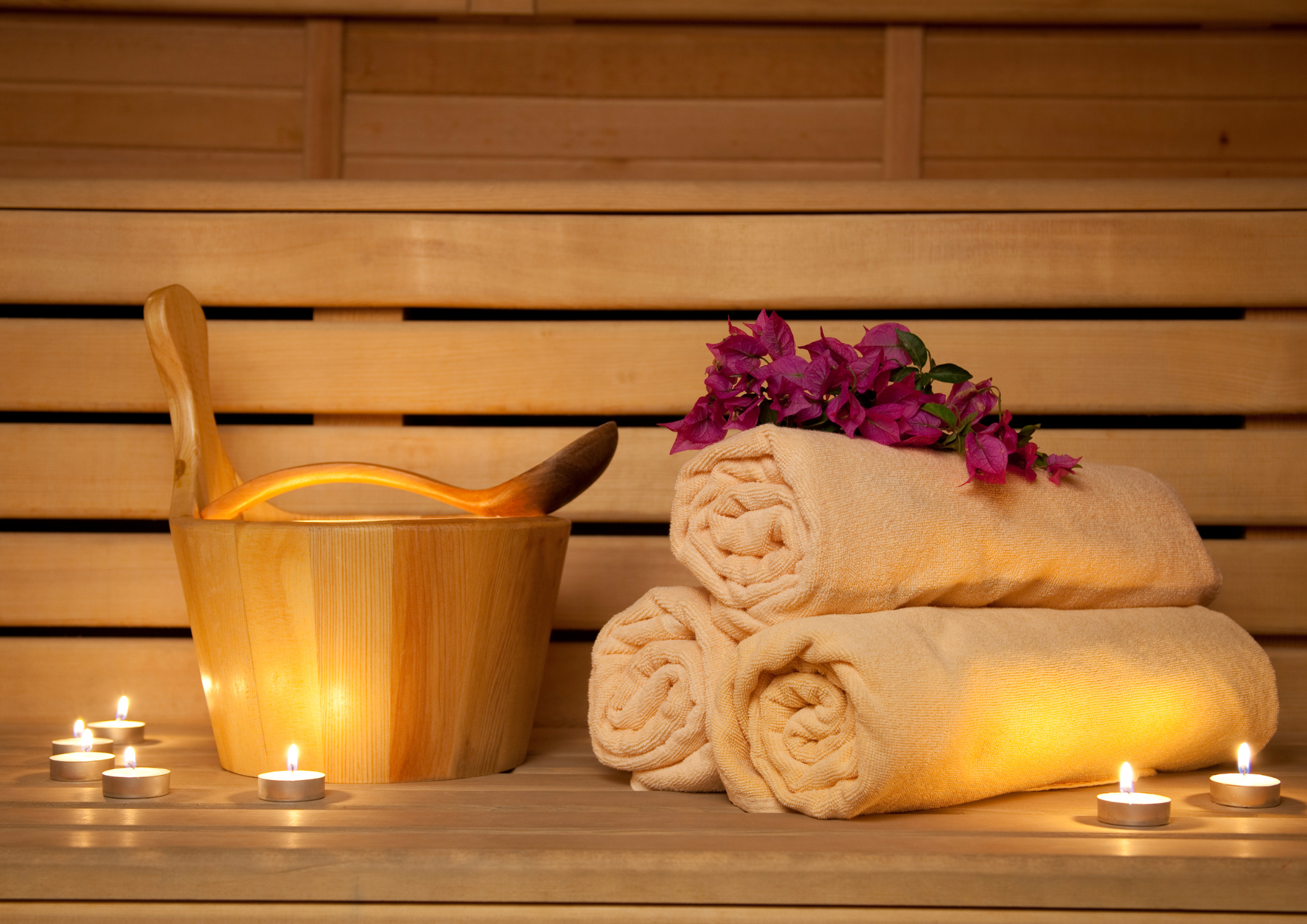 Spa setting with rolled towels, pink flowers, candles, and a wooden bucket on a wooden surface with candles around.