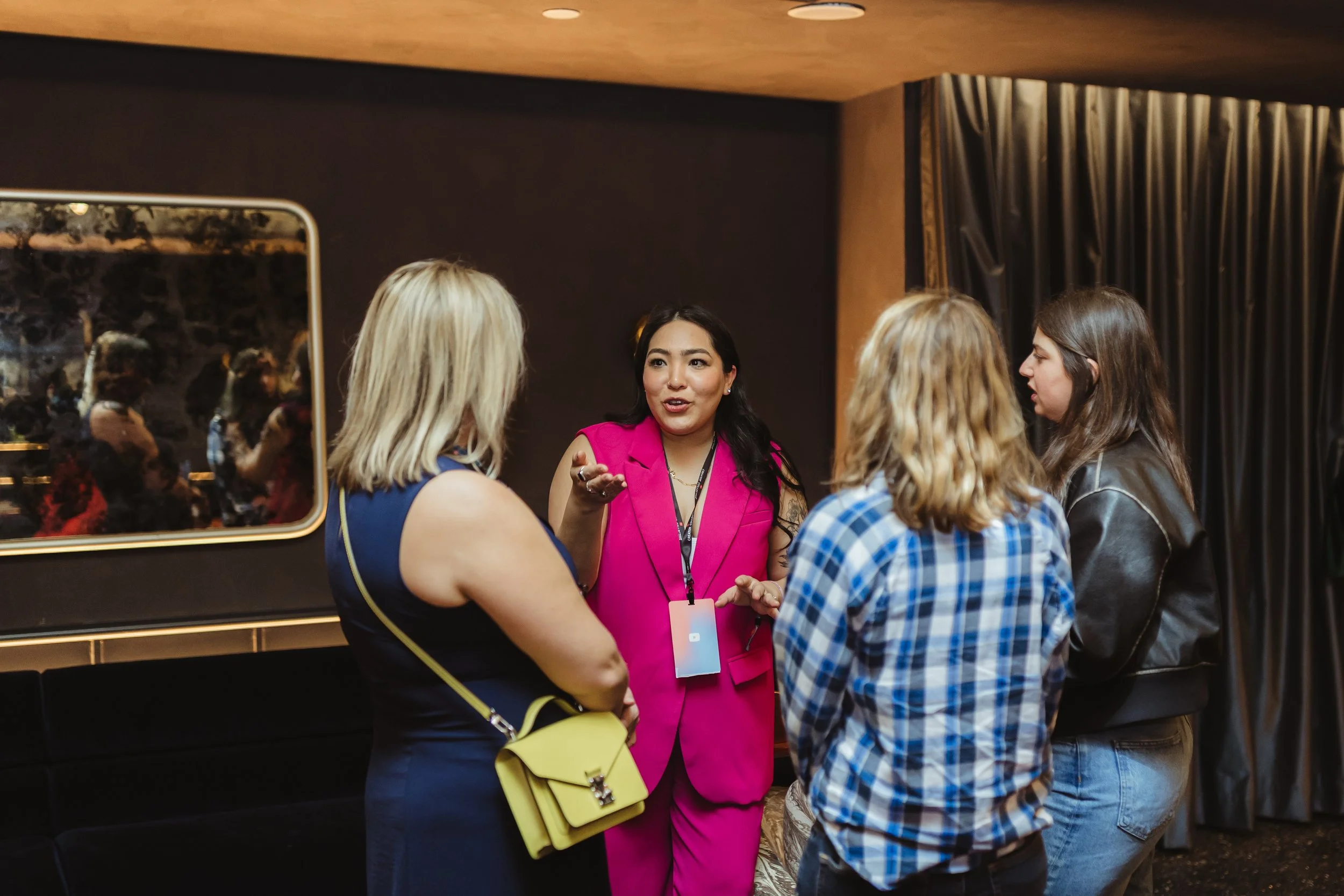 A woman in a pink suit speaking to three women in a dimly lit room with dark walls and curtains