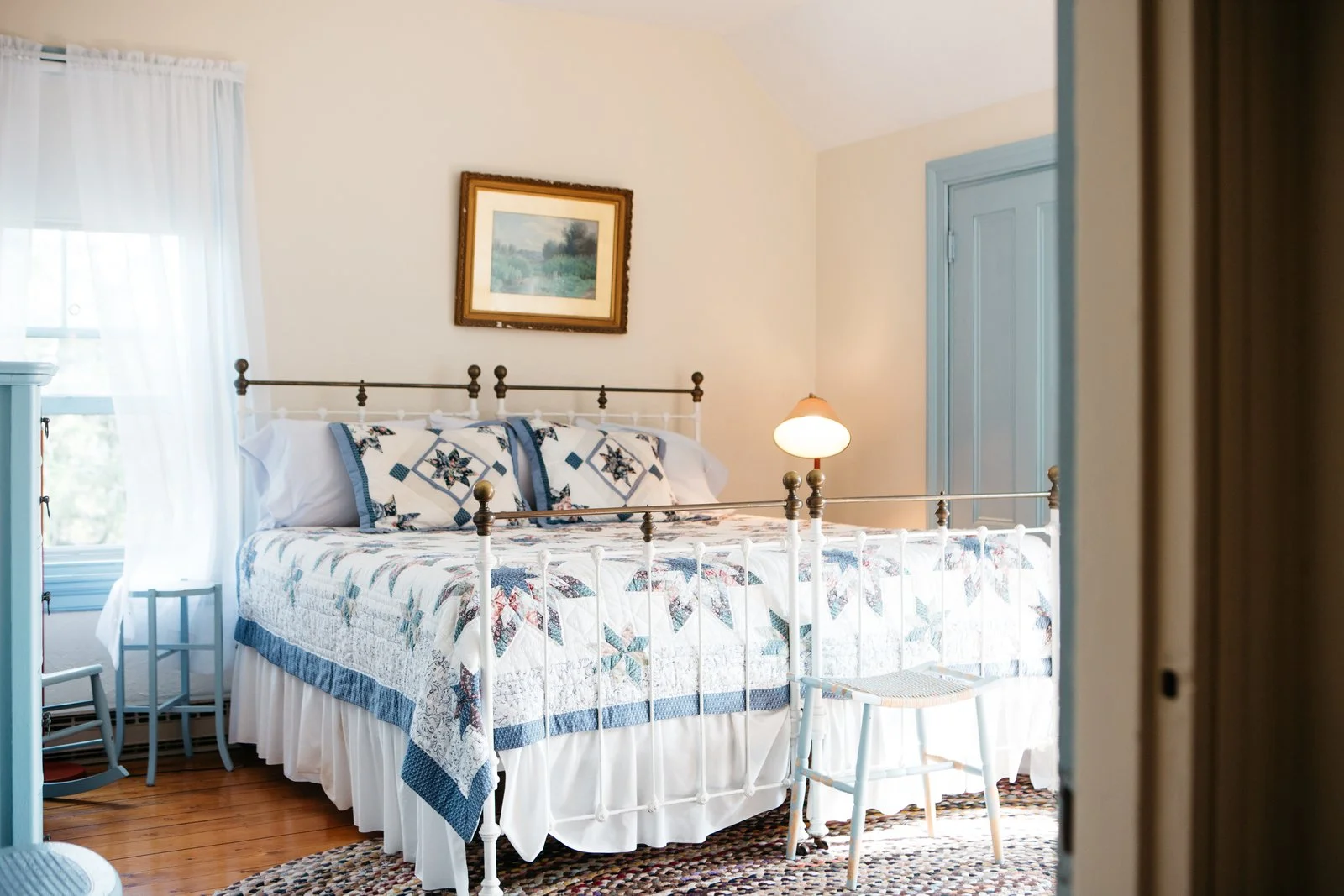 A cozy bedroom with a white iron bed, quilted bedspread, and matching pillows with blue accents. There is a wooden nightstand with a small lamp and a framed landscape painting on the wall. Natural light filters through sheer curtains on a window.