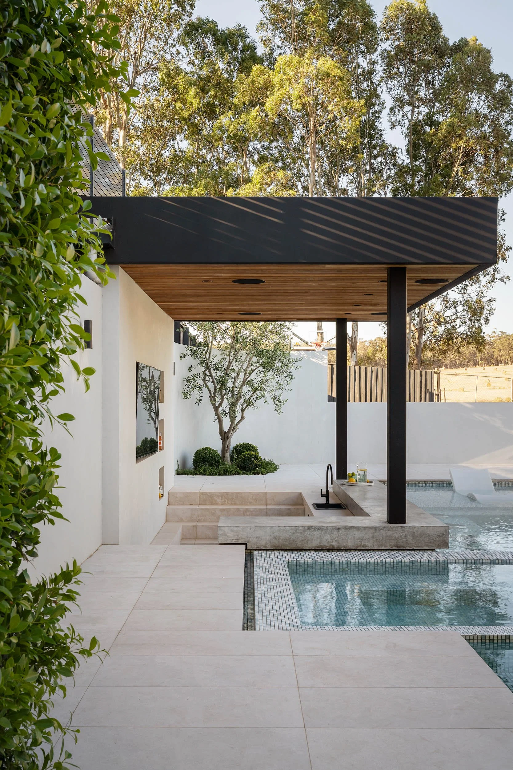 Modern backyard with a pool, outdoor kitchen, and shaded seating area, surrounded by greenery and trees.