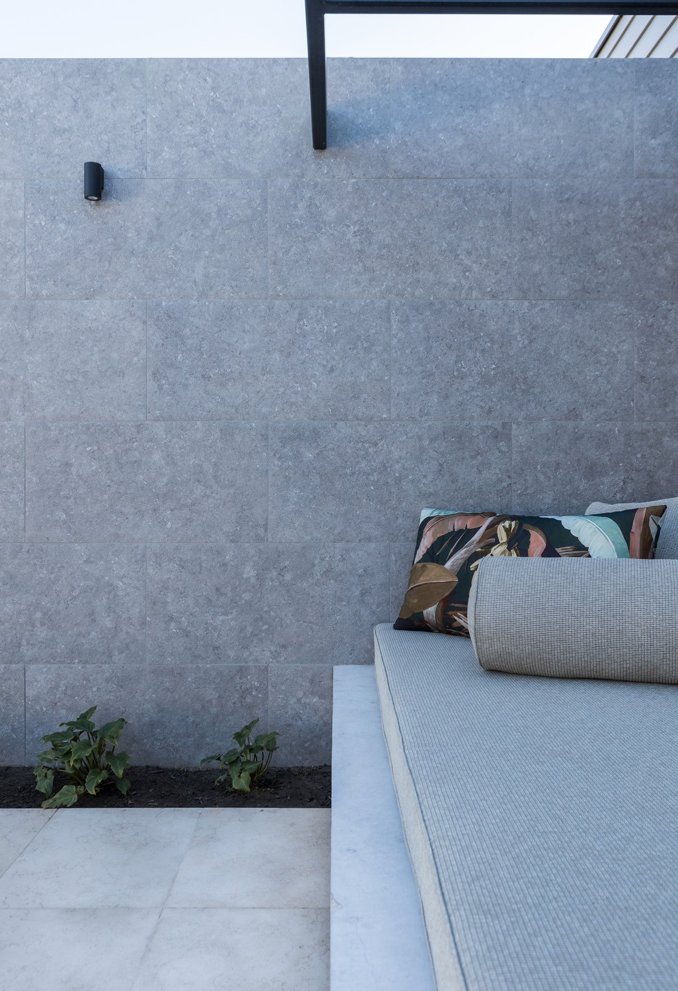 Outdoor gray stone wall with a bench featuring a cushion and decorative pillow, small plants, and a minimalist wall light.