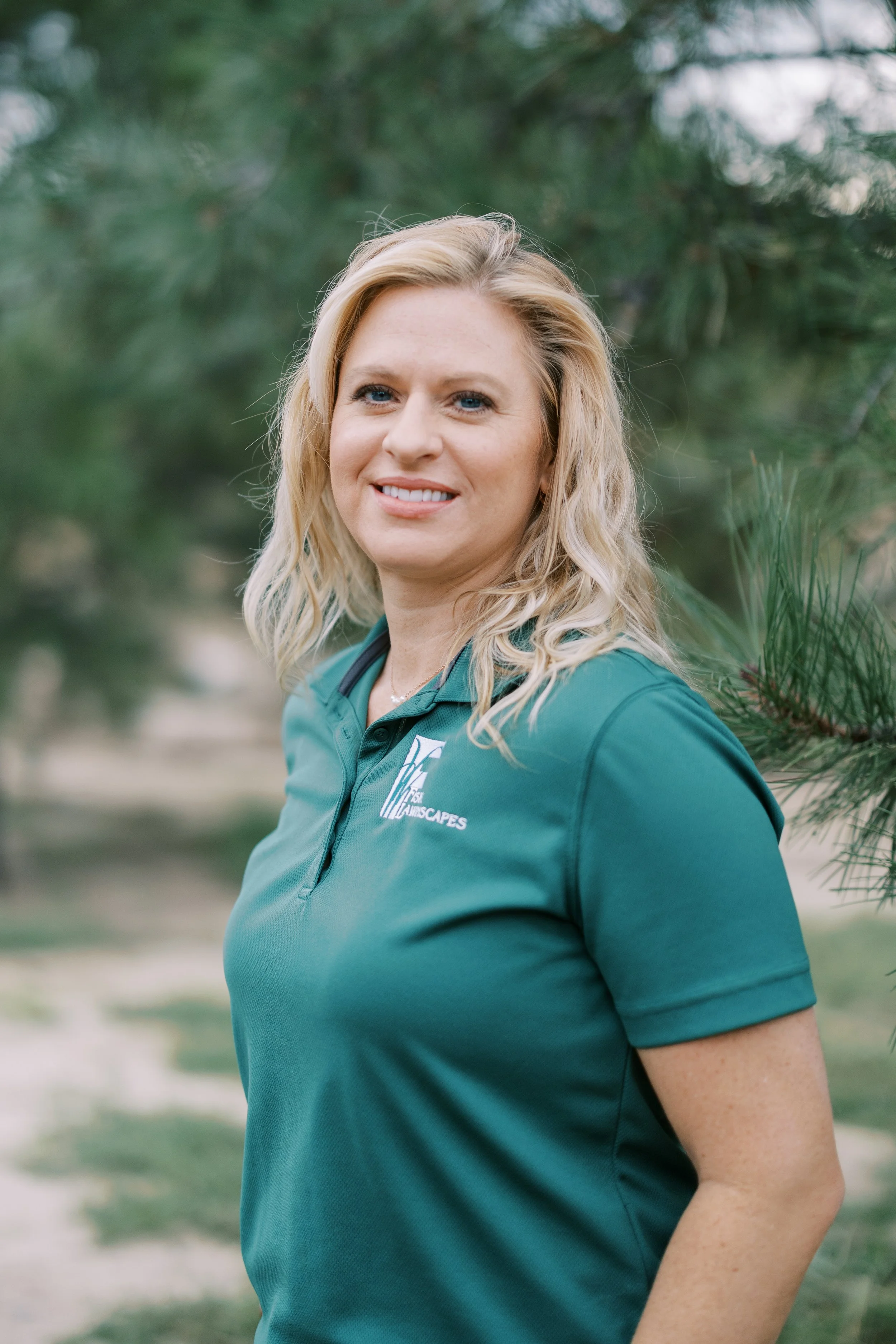 A woman with blonde hair wearing a black polo shirt with a logo that says 'Fisk Landscapes' outdoors during daytime, with trees and a body of water in the background.