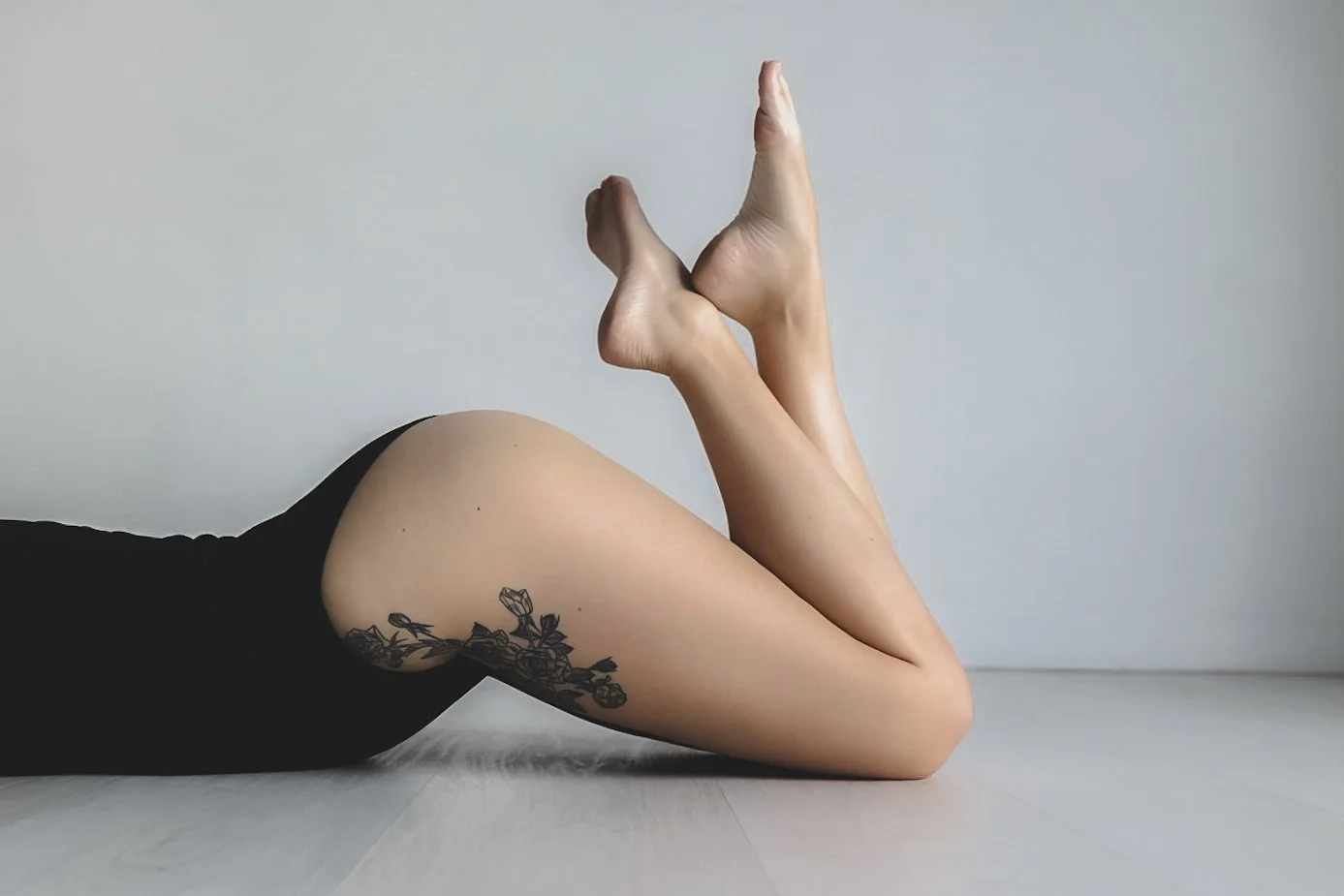Close-up of a person lying on the floor with their legs bent and crossed at the ankles, wearing black clothing, and showing a floral tattoo on their upper thigh.