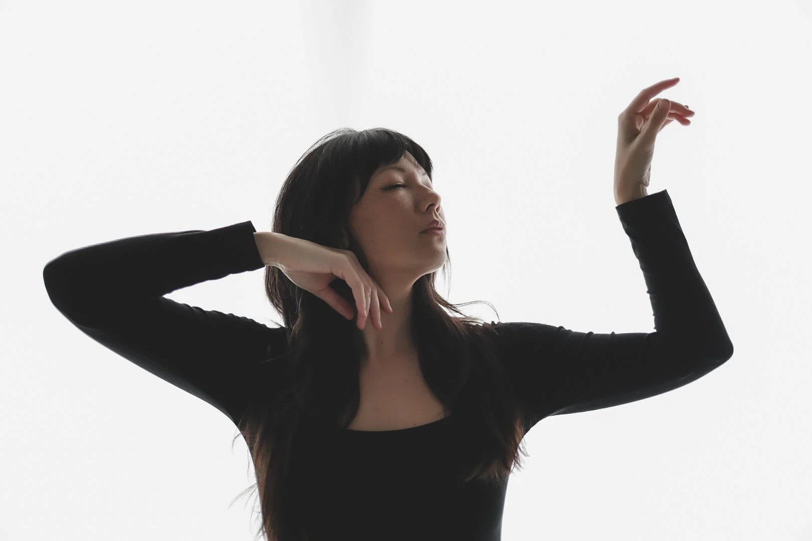 A woman with dark hair and a black long-sleeve top is posing with her eyes closed and her arms raised, one bent with her hand near her face and the other extended above her head against a plain white background.