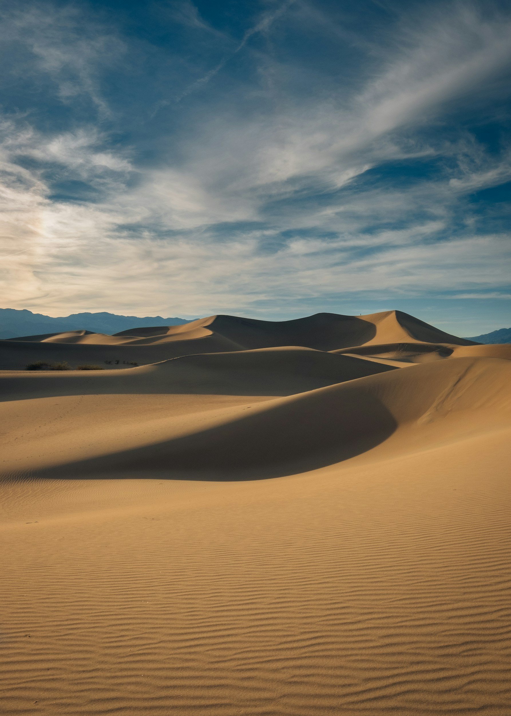 Image of desert sand dunes under a partly cloudy sky with mountains in the background.