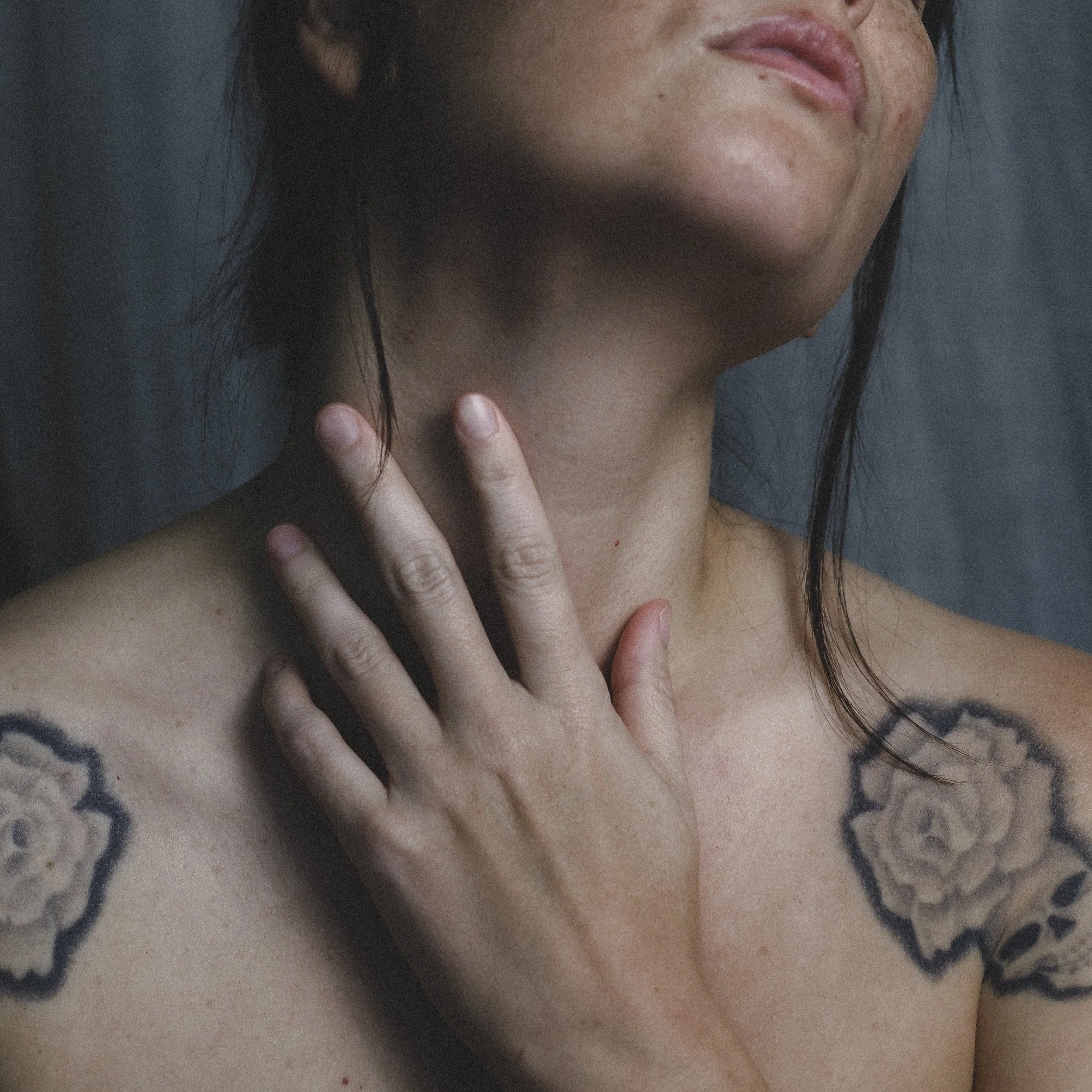 A close-up of a woman's shoulder, neck, and hand with tattoos, her face partially visible, with a dark background.