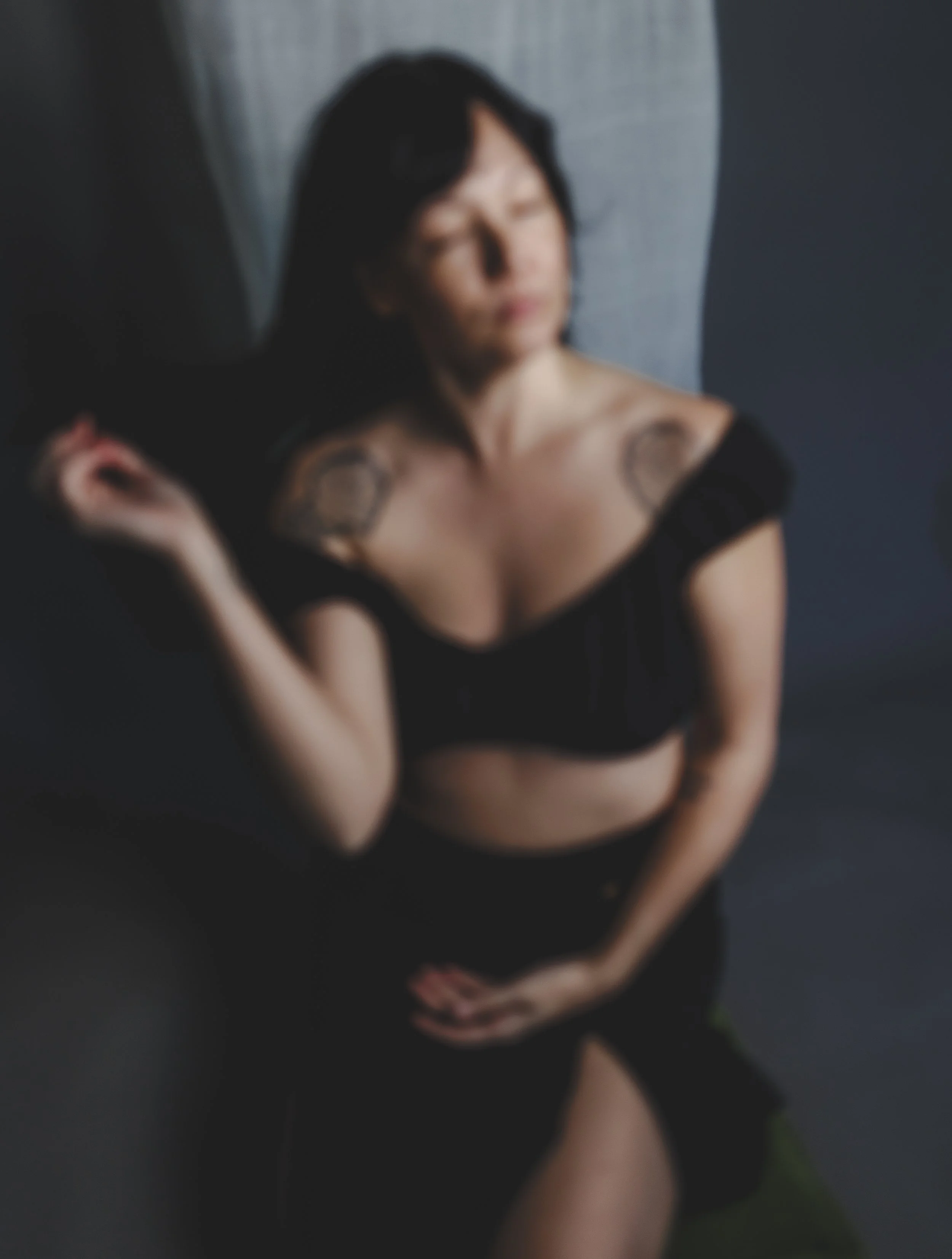 A woman with dark hair, wearing a black top and shorts, is sitting against a dark background with her eyes closed and head tilted back.