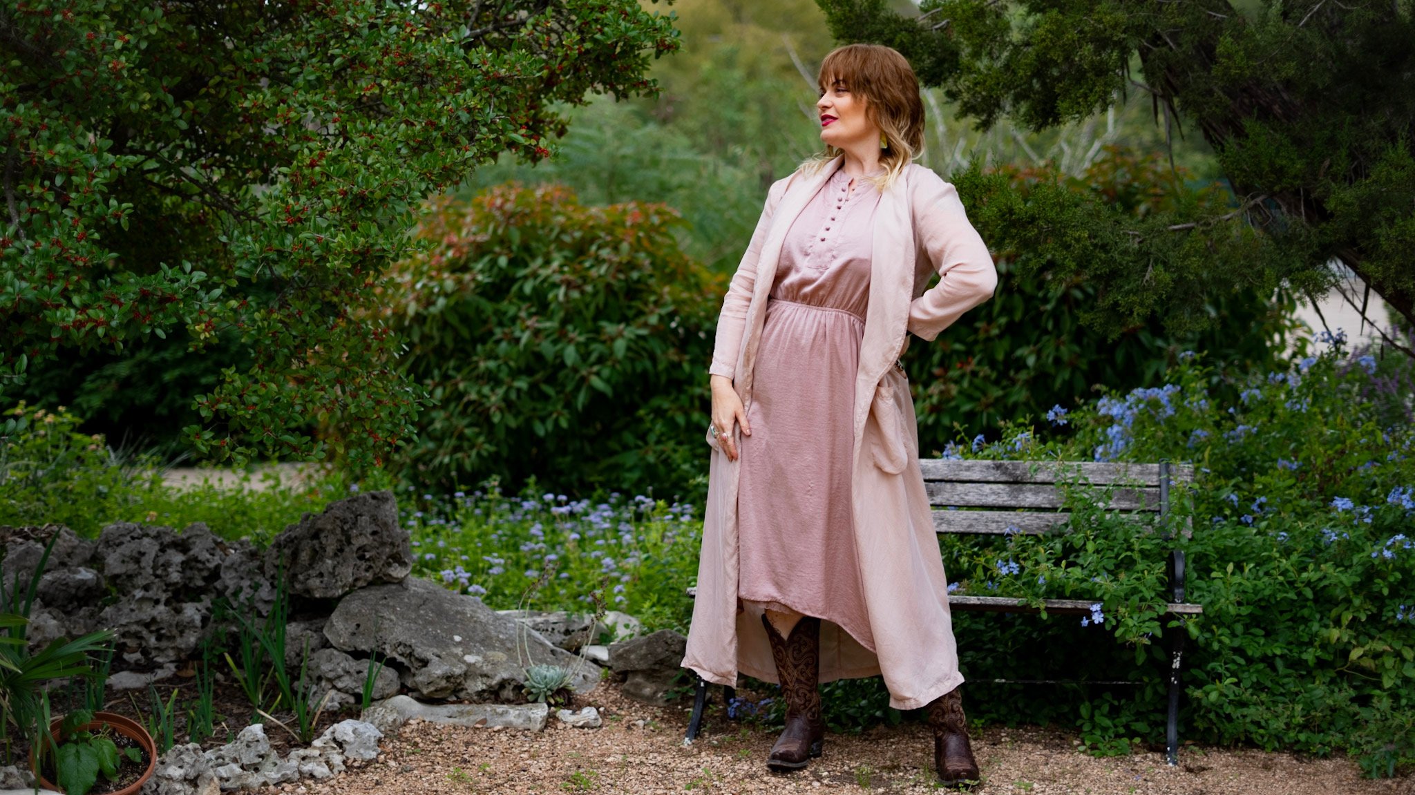 Woman in a pink dress and matching coat standing outdoors in a garden, with her hands on her hips, surrounded by greenery, bushes, and flowers, with a bench in the background.