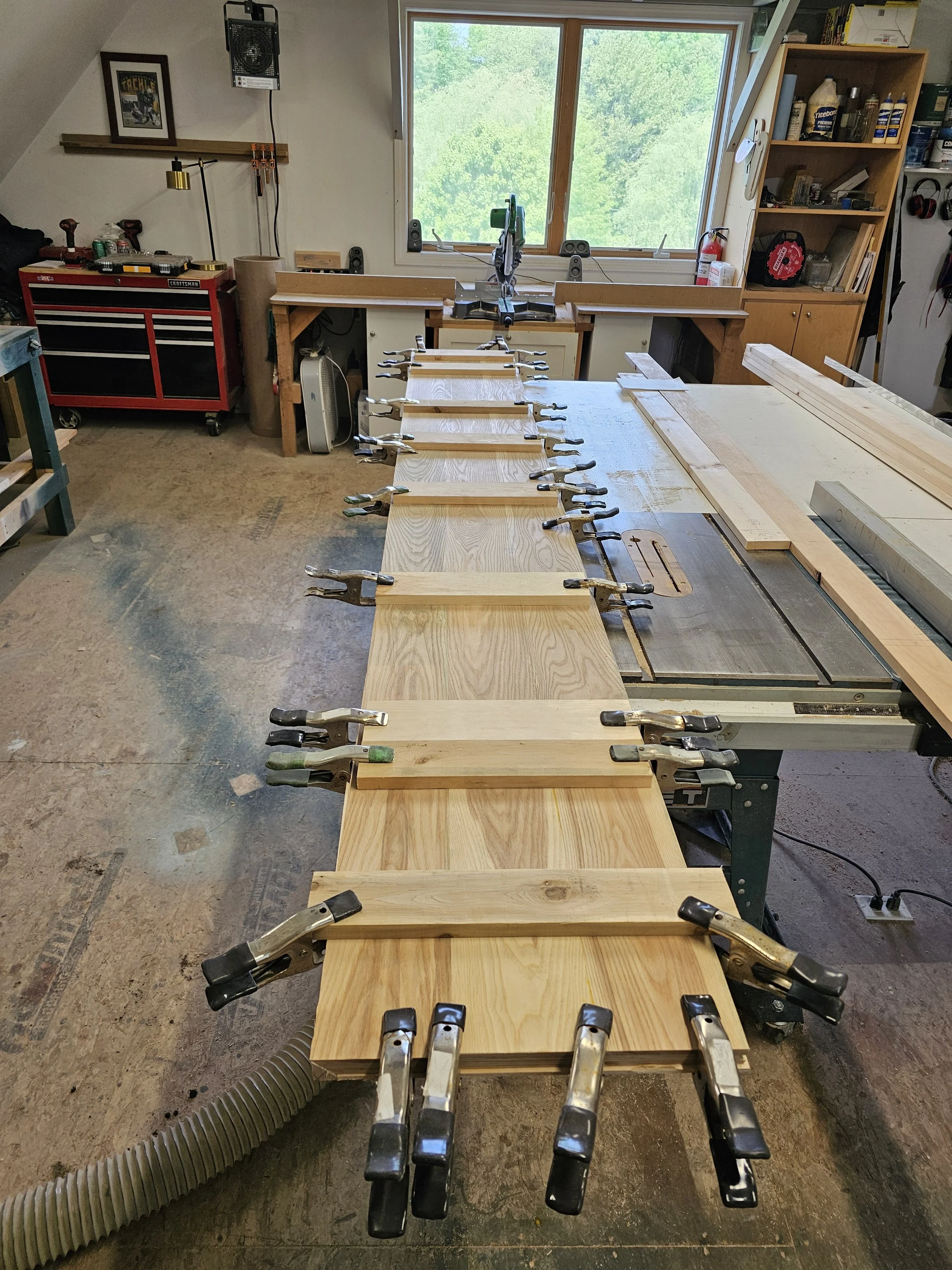 A woodworking shop with a workbench holding several pieces of wood clamped in place. In the background, there is a saw and a window showing green trees outside. The room contains tools, storage cabinets, and woodworking equipment.