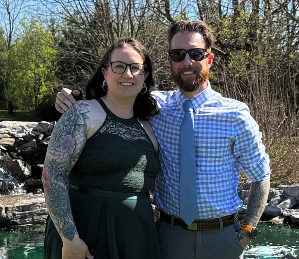 A smiling woman and man standing outdoors near a small waterfall, with trees in the background. The woman has tattoos on her arm and is wearing glasses and a black dress. The man has a beard, is wearing sunglasses, a checkered shirt with rolled-up sleeves, a blue tie, and dark pants.