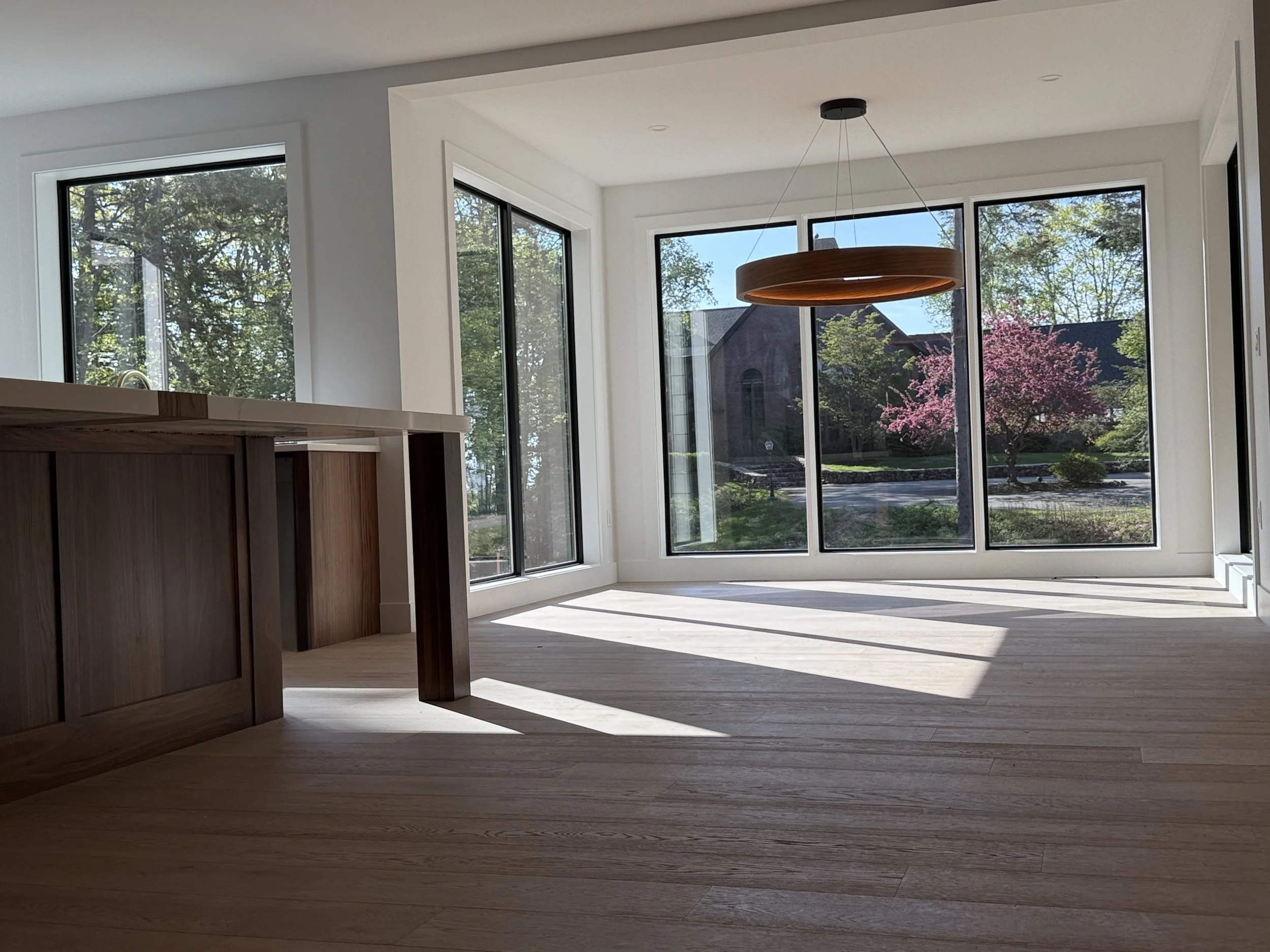 Bright living room with large windows showing trees and a pink flowering tree outside, wooden floor, and a modern circular wooden chandelier hanging from the ceiling.