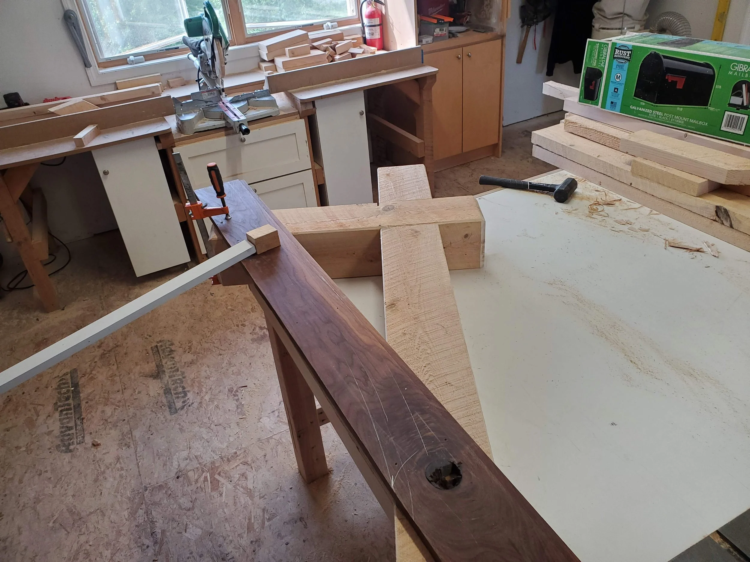 A woodworking workshop with a workbench, a circular saw, and several pieces of wood. There's a window with a fire extinguisher on the wall, and a box of galvanized steel mailbox parts on a stacked set of wood boards.