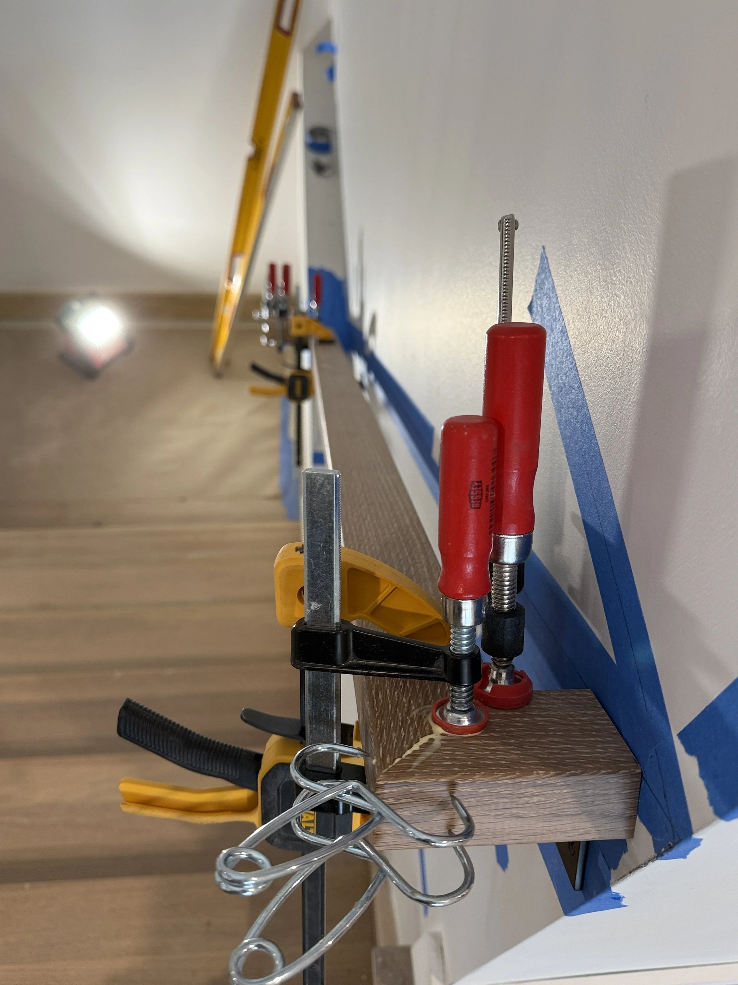Tools such as a yellow clamp, a screwdriver with a red handle, a measuring scale, and a black clip are attached to the edge of a wooden surface, with blue painter's tape on the wall for marking.