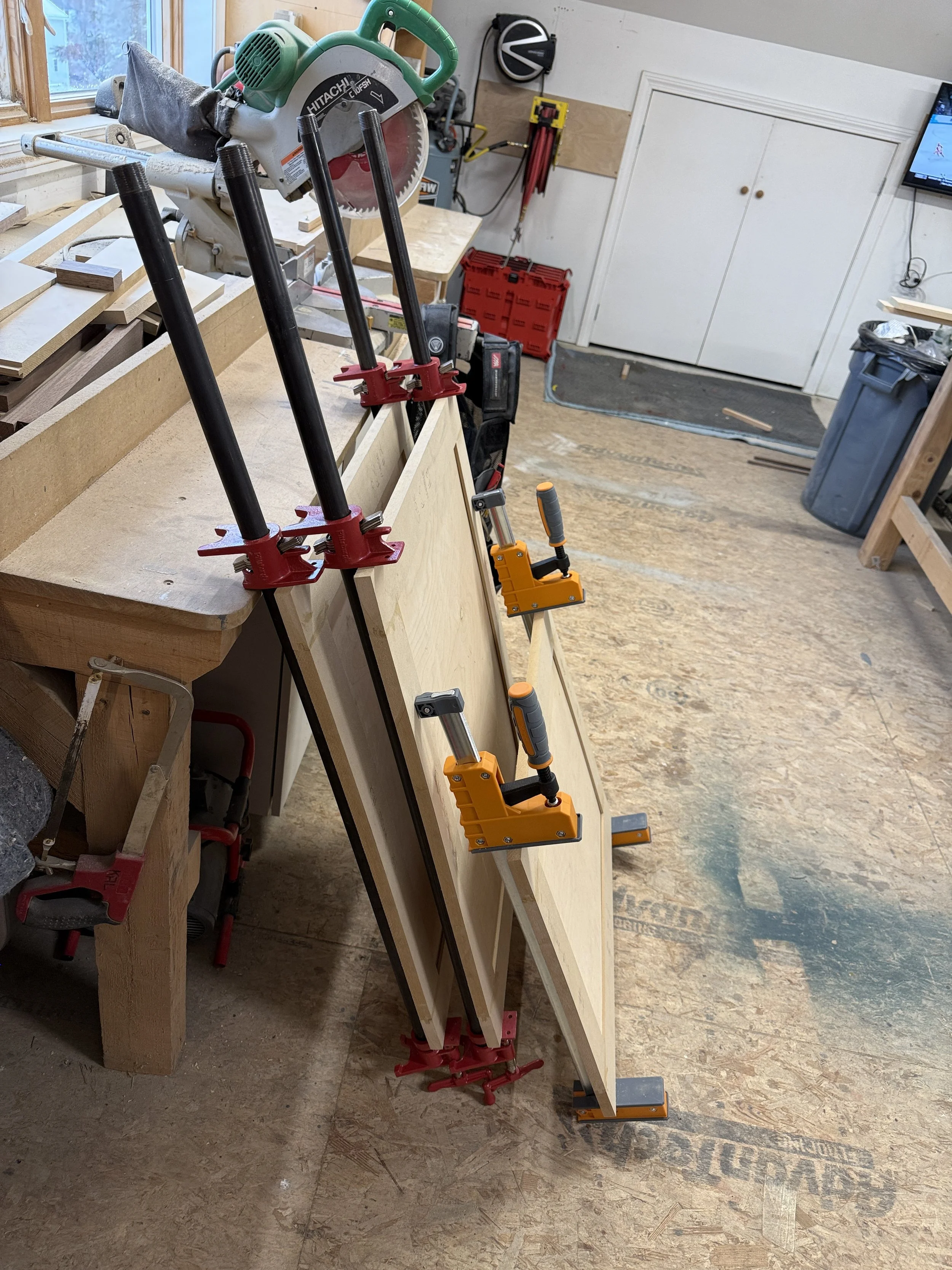 A woodworking workshop with a bandsaw, clamps, and wood panels on a workbench. There is a TV on the wall, a trash can, and various tools and equipment in the background.