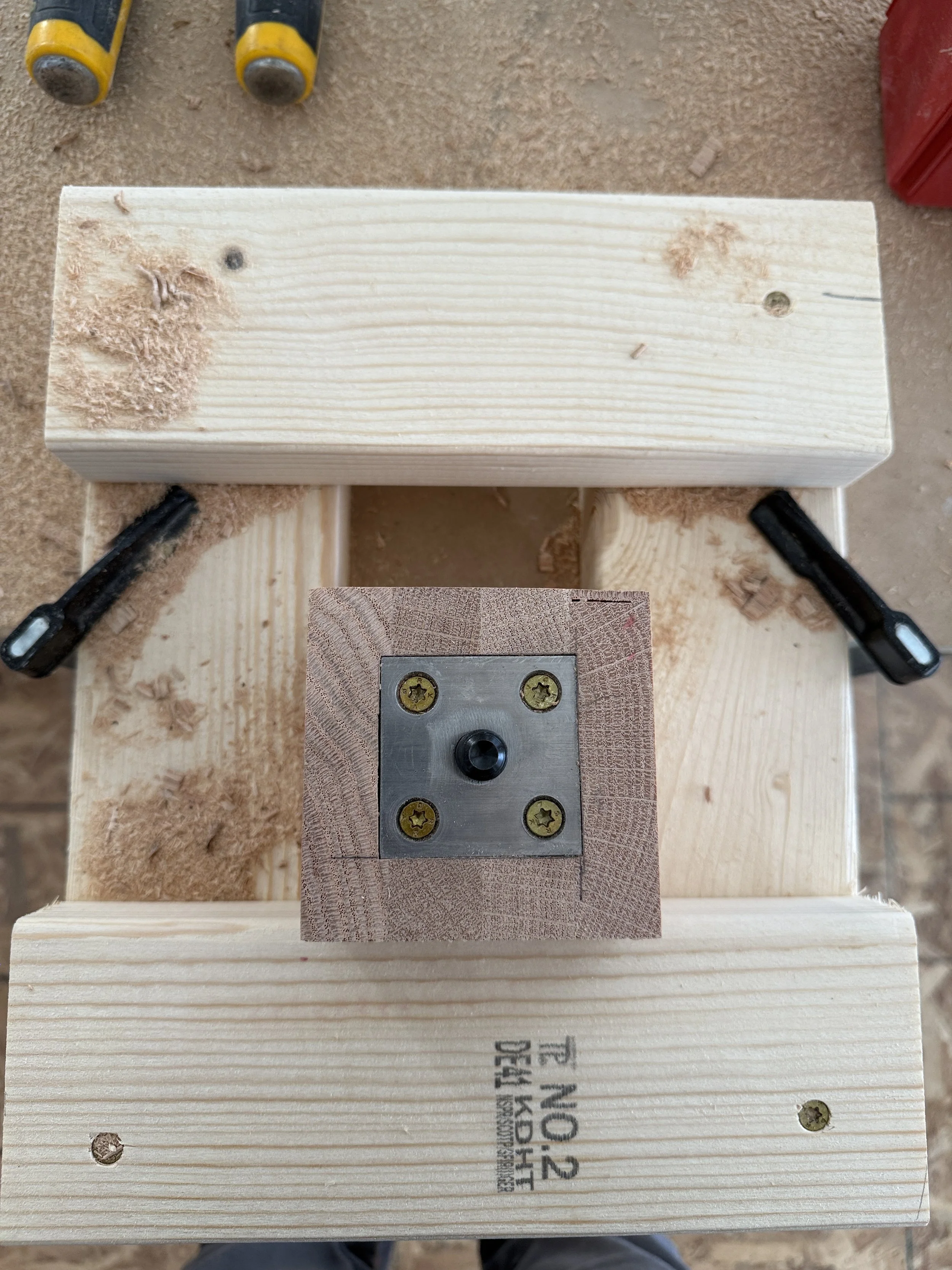 Close-up view of a woodworking project with a wooden block attached to a larger piece, secured with screws and bolts, on a workbench with tools and sawdust.