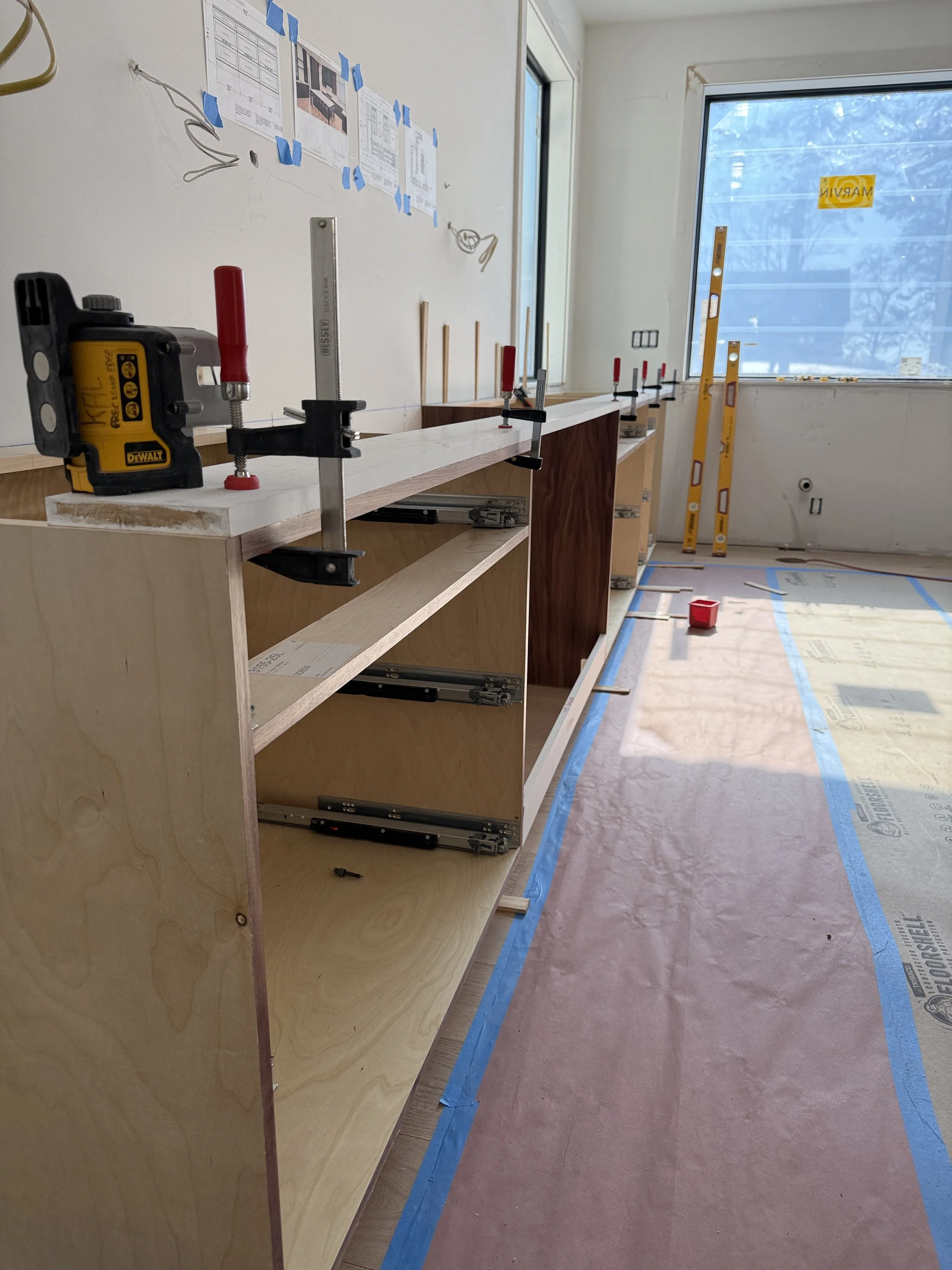 Unfinished wooden cabinet with clamps holding a piece of white wood in place, inside a room under construction. There are tools, leveling instruments, and taped documents on the walls, with large windows allowing natural light.
