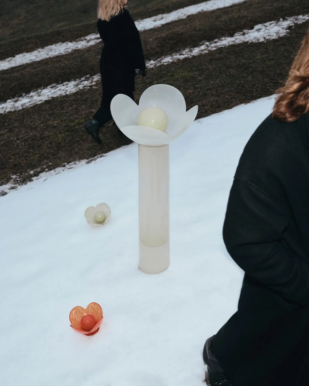 The flower-shaped Lucky Akebia Floor Lamp and two Lucky Akebia Desk Lamps in the snow with the designers, Anna and Sergii Baierzdorf, walking by.