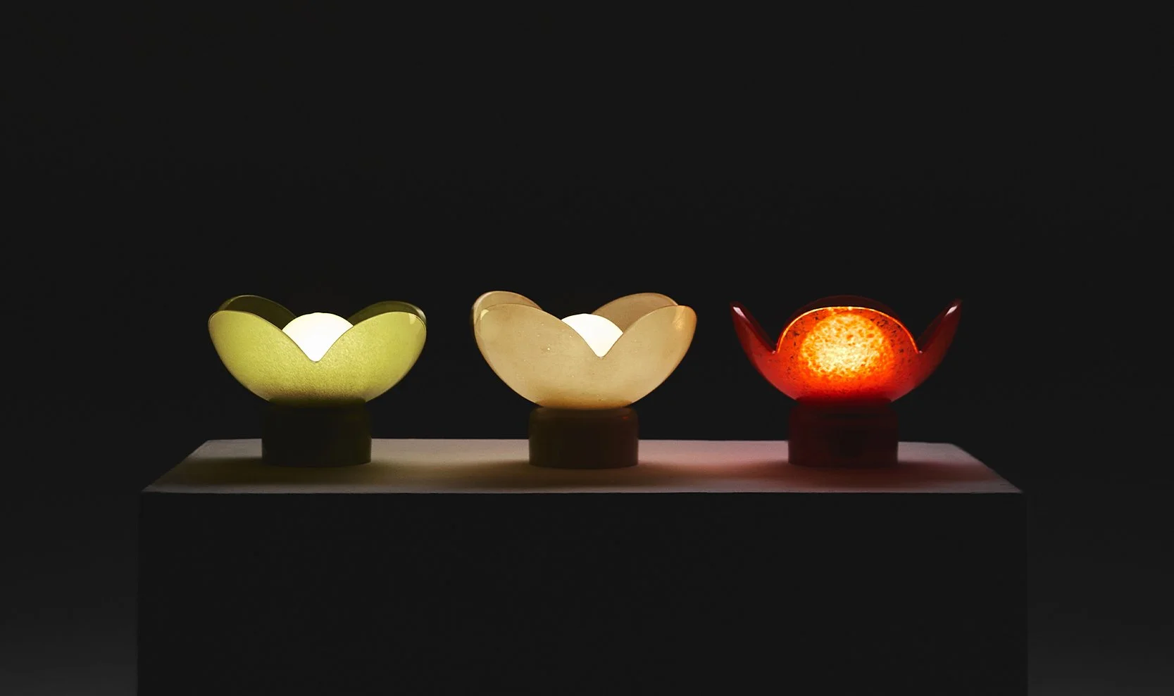The green, red and beige flower-shaped Lucky Akebia Desk Lamps by Furn Object lit up side by side in a dark room.